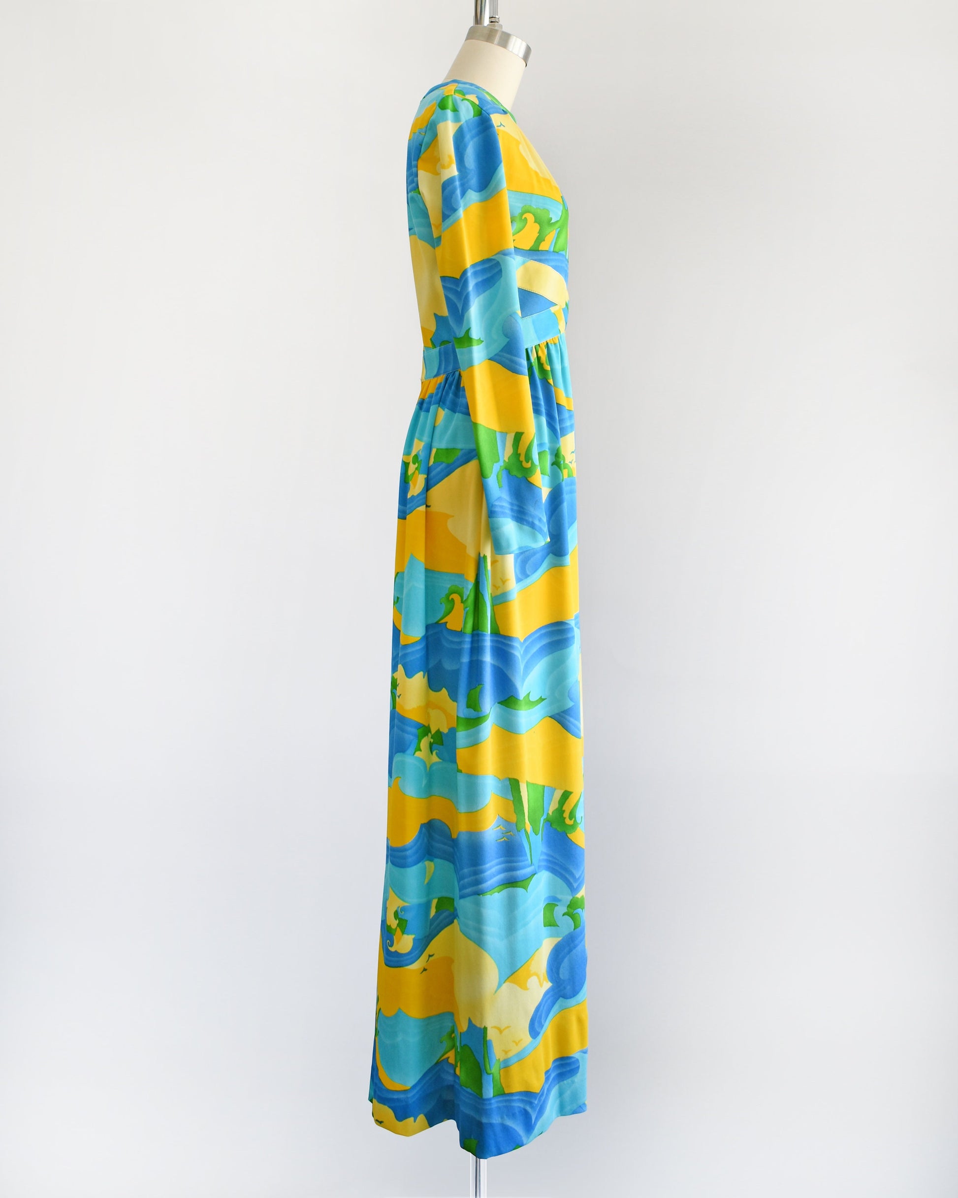 Side view of a vintage 70s maxi dress with a blue, yellow, and green ocean landscape print that has sea, sand, and sky, with greenery in the back and birds in the sky. The dress has long sleeves and is on a dress form.