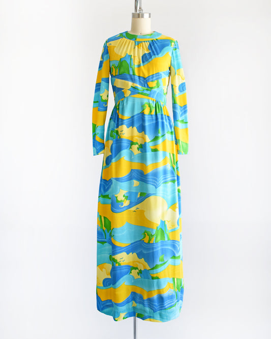 A vintage 70s maxi dress with a blue, yellow, and green ocean landscape print that has sea, sand, and sky, with greenery in the back and birds in the sky. The dress has long sleeves and is on a dress form.