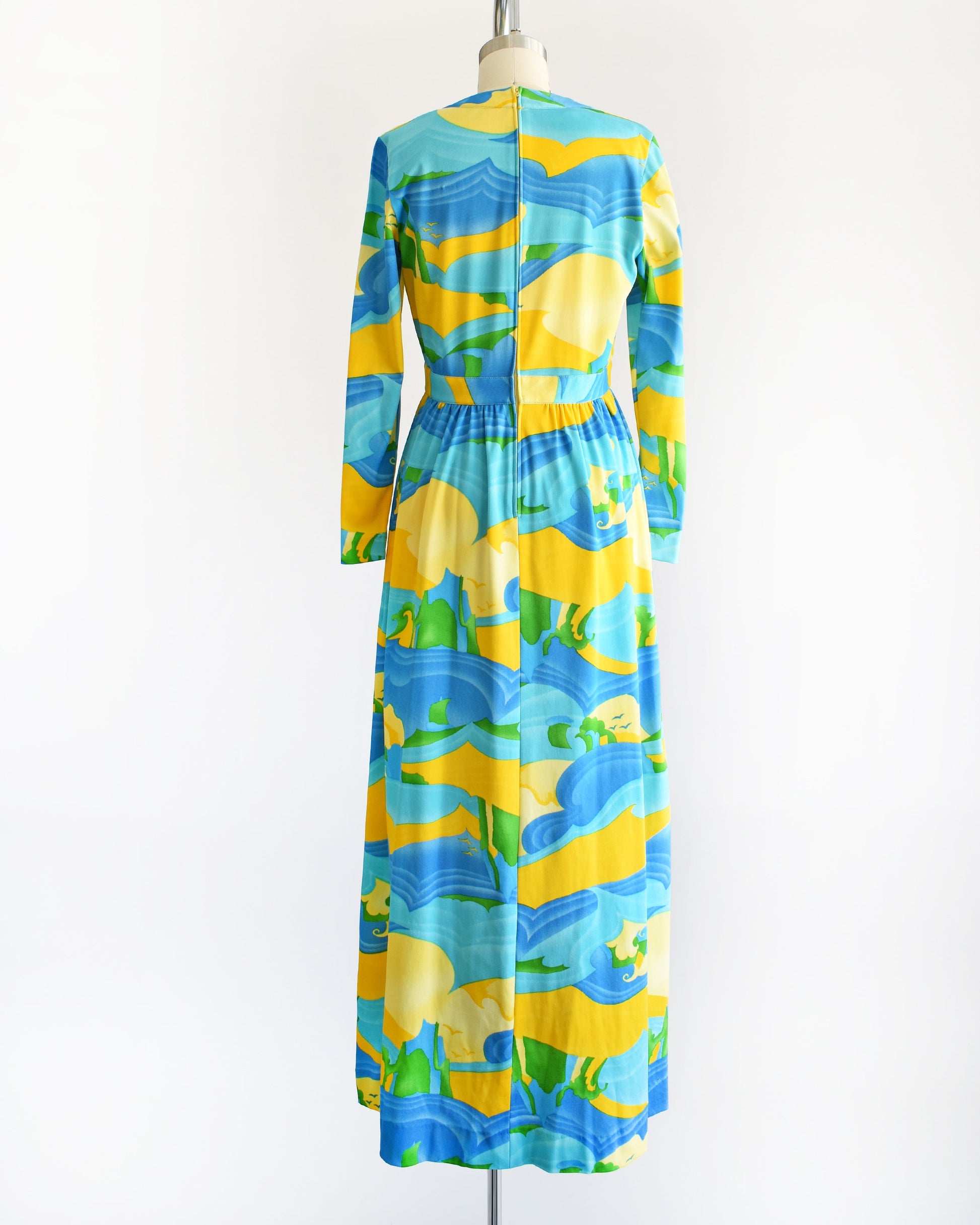Back view of a vintage 70s maxi dress with a blue, yellow, and green ocean landscape print that has sea, sand, and sky, with greenery in the back and birds in the sky. The dress has long sleeves and is on a dress form.