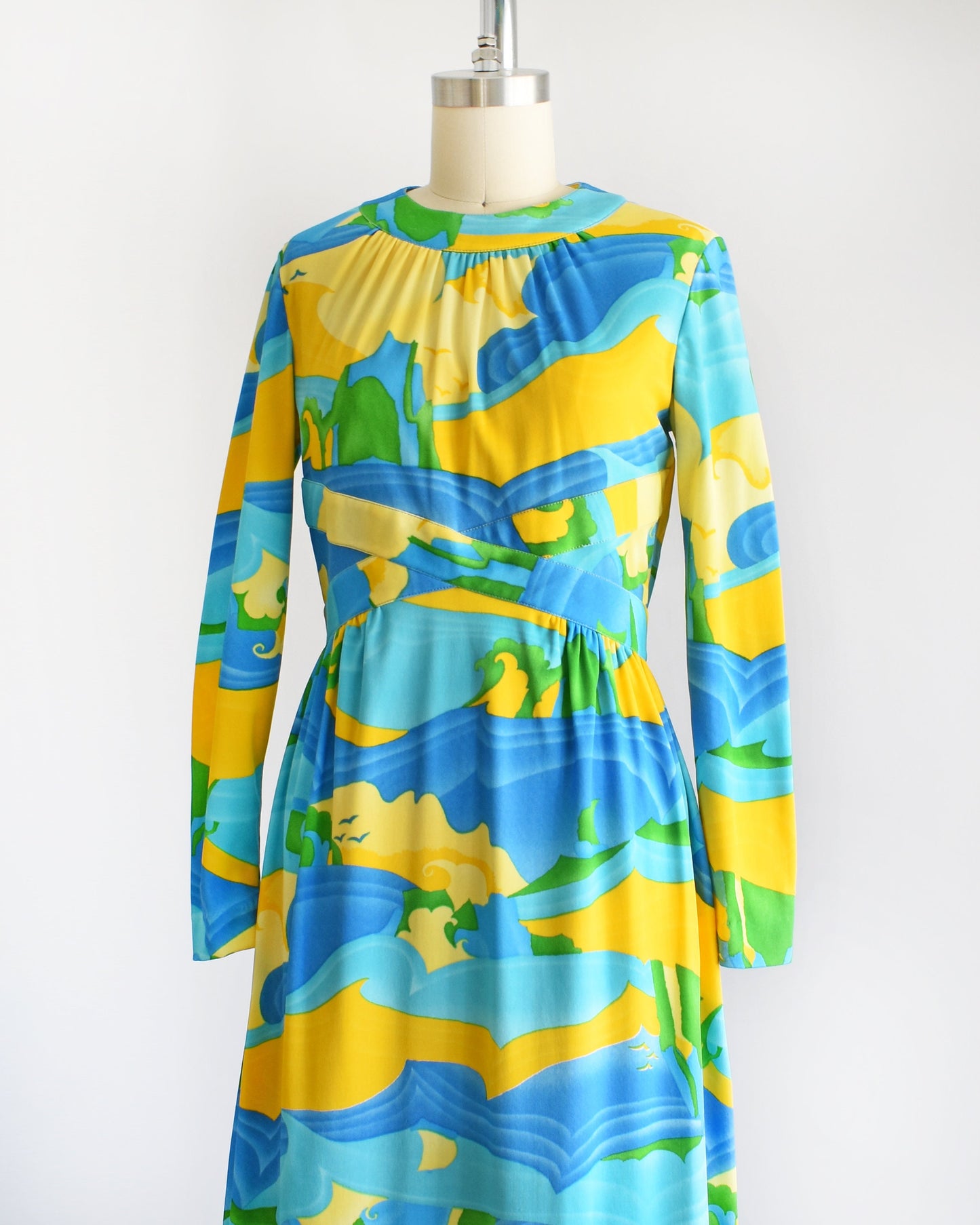 Side front view of a vintage 70s maxi dress with a blue, yellow, and green ocean landscape print that has sea, sand, and sky, with greenery in the back and birds in the sky. The dress has long sleeves and is on a dress form.