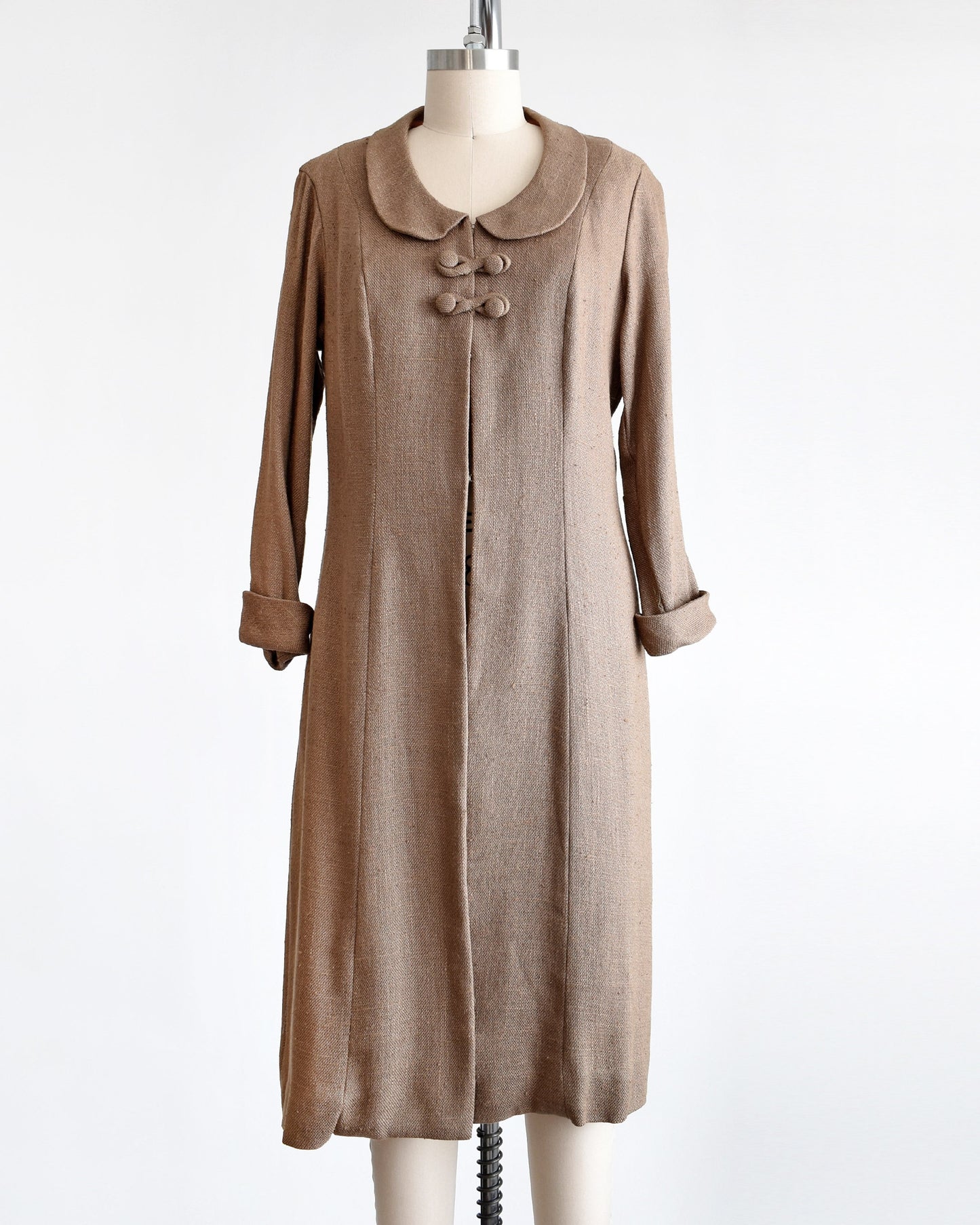 A vintage late 50s early light brown wool swing coat. The coat has a rounded Peter Pan style collar, four buttons underneath and cuffed long sleeves. The coat is modeled on a dress form.