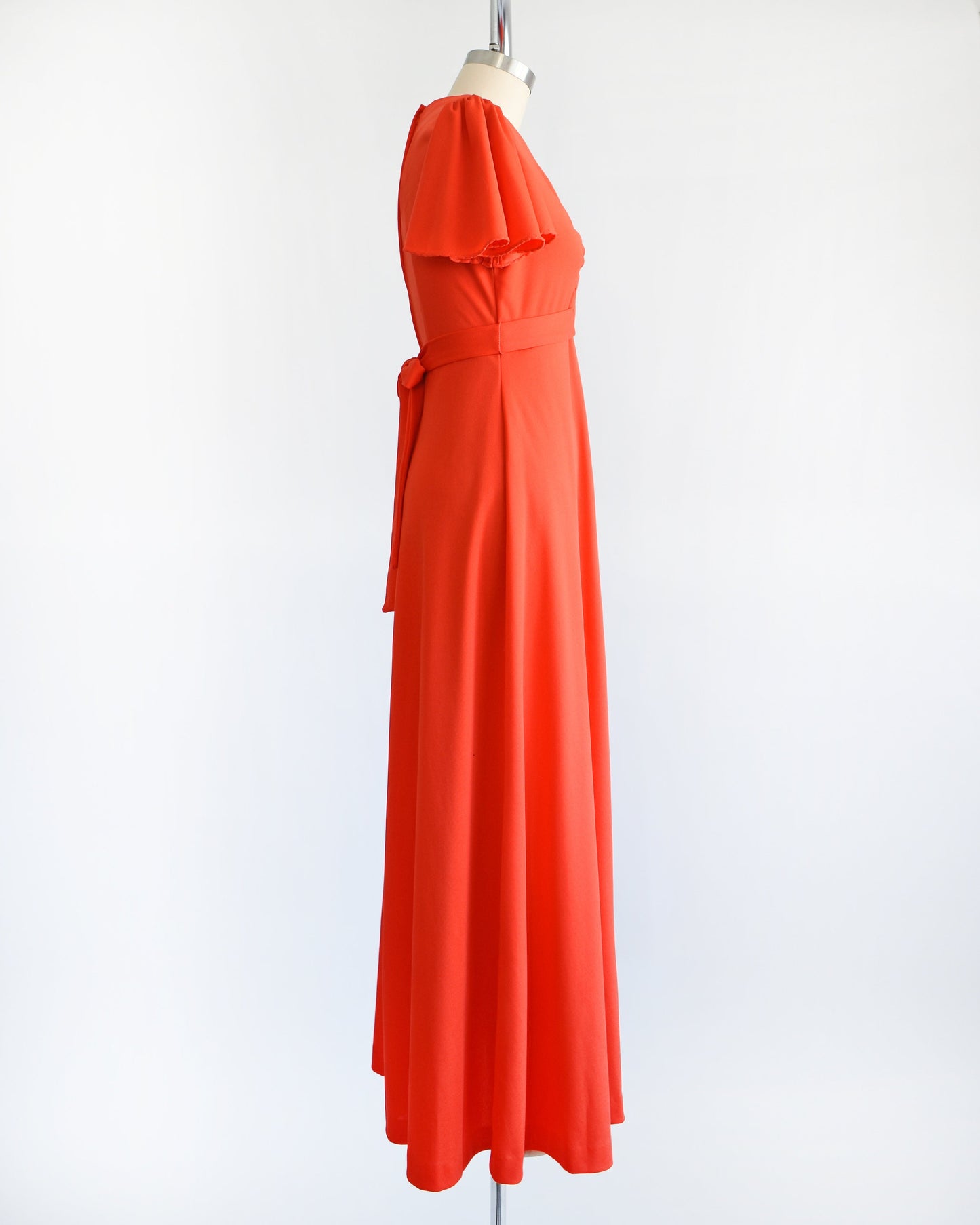 Side view of a vintage 70s red orange maxi dress which features short sleeves, v-neckline with silver studs and rhinestones underneath. Empire waist and long flowing skirt. The dress is displayed on a dress form.