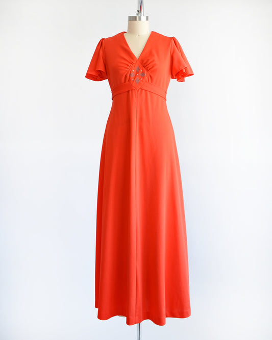 A vintage 70s red orange maxi dress which features short sleeves, v-neckline with silver studs and rhinestones underneath. Empire waist and long flowing skirt. The dress is displayed on a dress form.