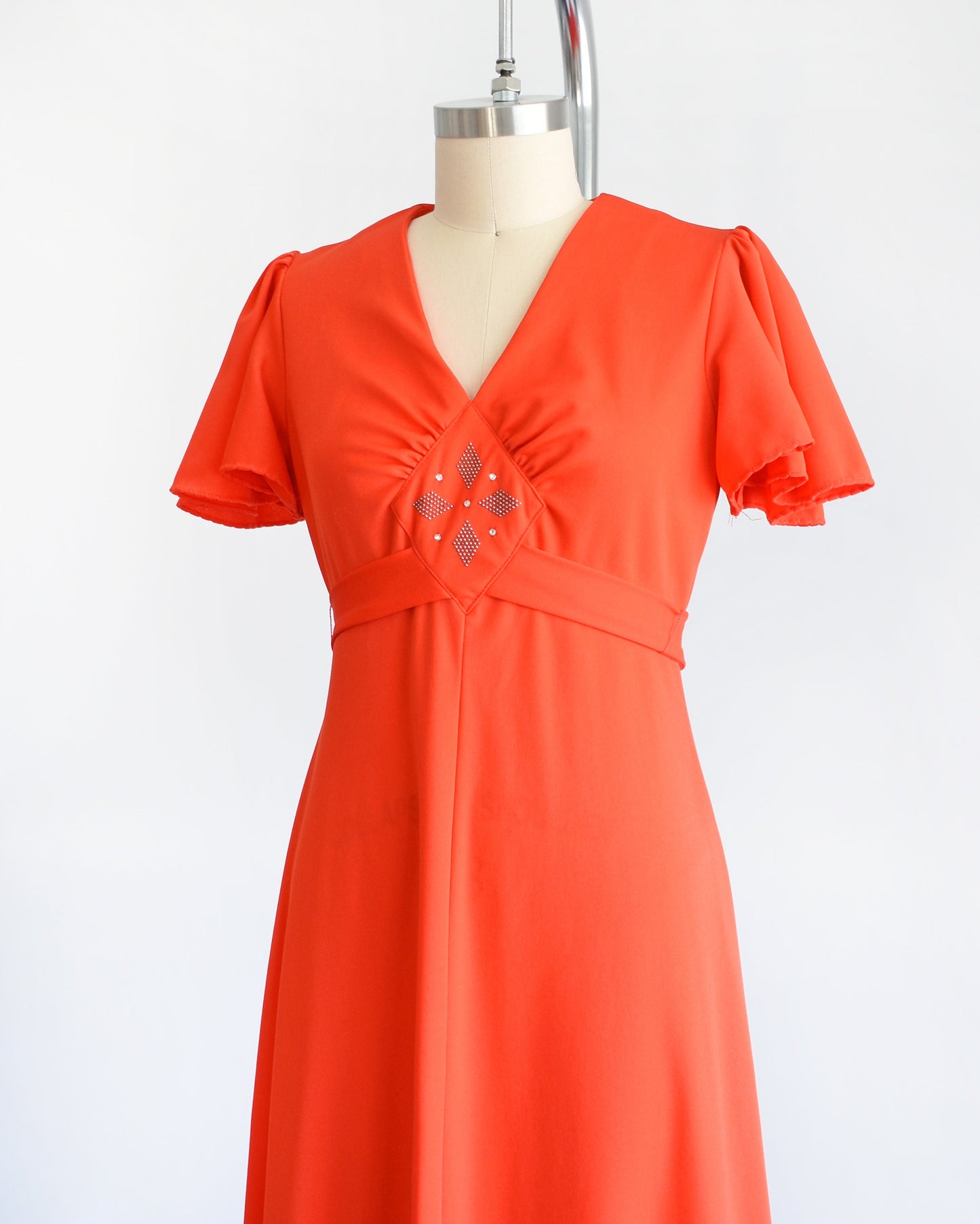 Side front view of a vintage 70s red orange maxi dress which features short sleeves, v-neckline with silver studs and rhinestones underneath. Empire waist and long flowing skirt. The dress is displayed on a dress form.