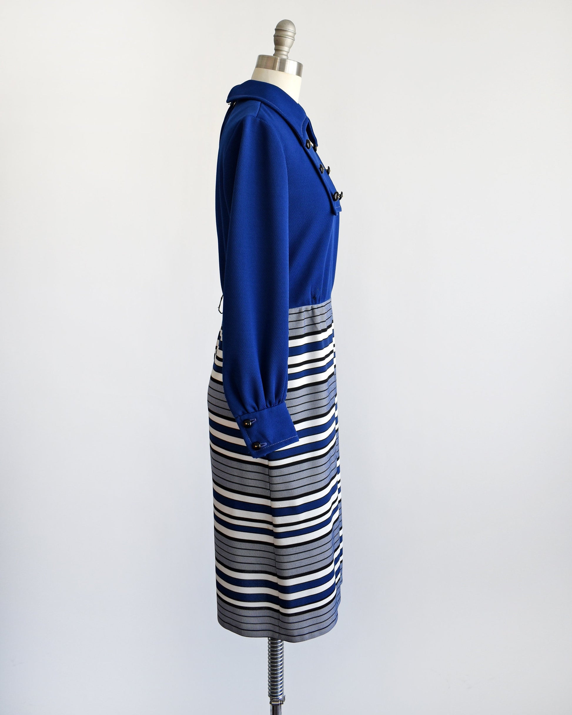 Side view of a vintage 60s mod dress on a dress form. The dress has a dark blue bodice with a pointed collar and six decorative buttons. Long sleeves. Blue, black, and white horizontal striped skirt with a large pleat on the front.