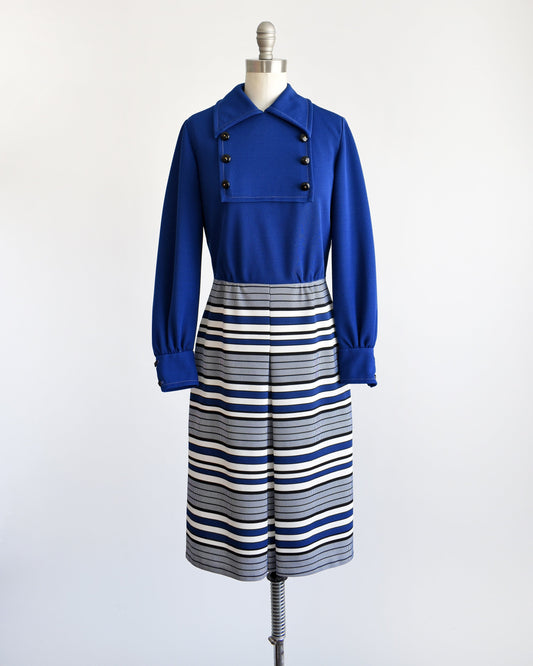 A vintage 60s mod dress on a dress form. The dress has a dark blue bodice with a pointed collar and six decorative buttons in two rows of threes. Long sleeves. Blue, black, and white horizontal striped skirt with a large pleat on the front.