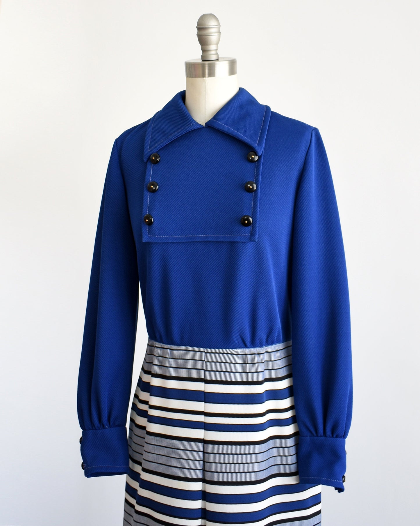 Side front view of a vintage 60s mod dress on a dress form. The dress has a dark blue bodice with a pointed collar and six decorative buttons in two rows of threes. Long sleeves. Blue, black, and white horizontal striped skirt with a front pleat.