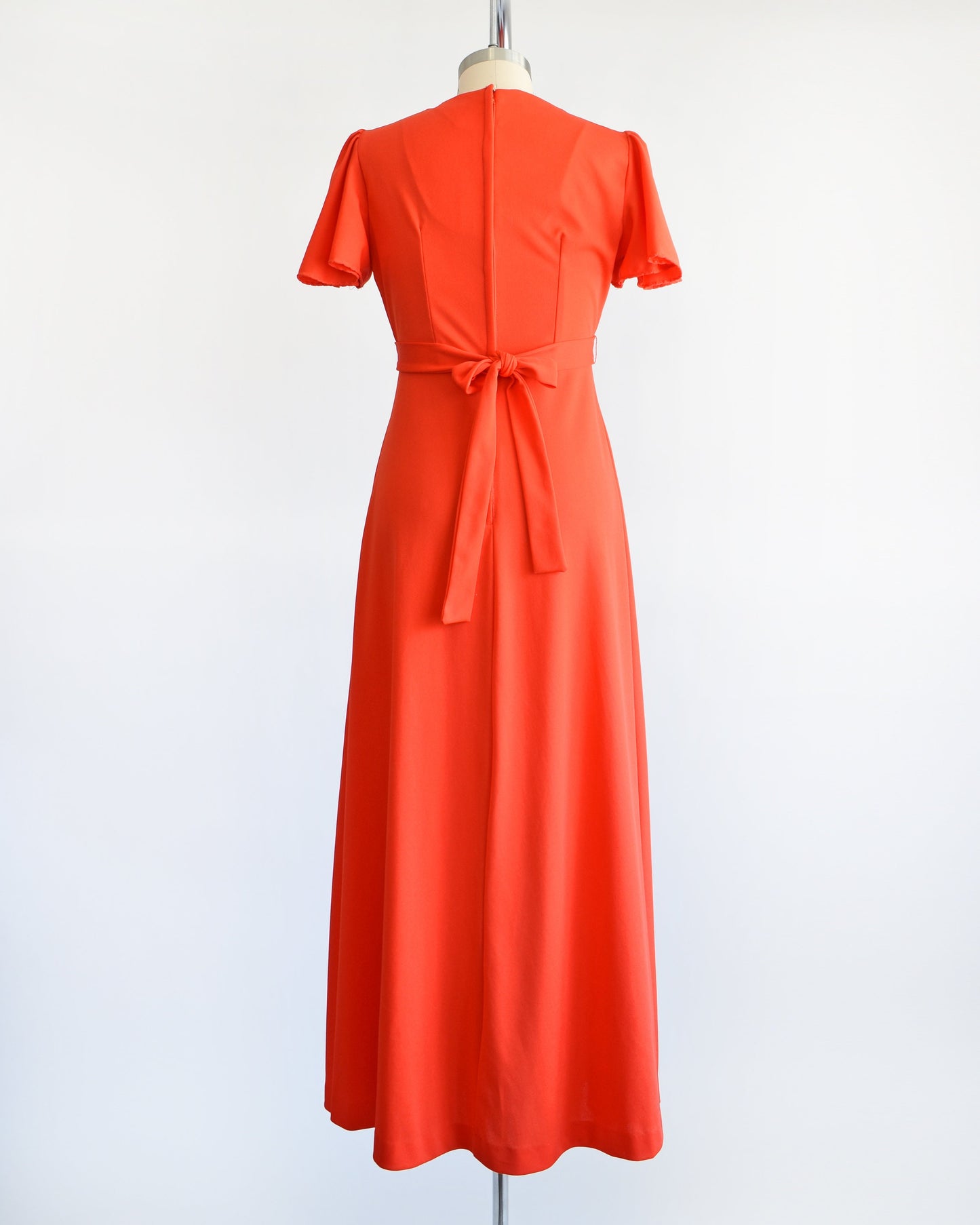Back view of a vintage 70s red orange maxi dress which features short sleeves, and empire waist with ties in the back, and long flowing skirt. The dress is displayed on a dress form.
