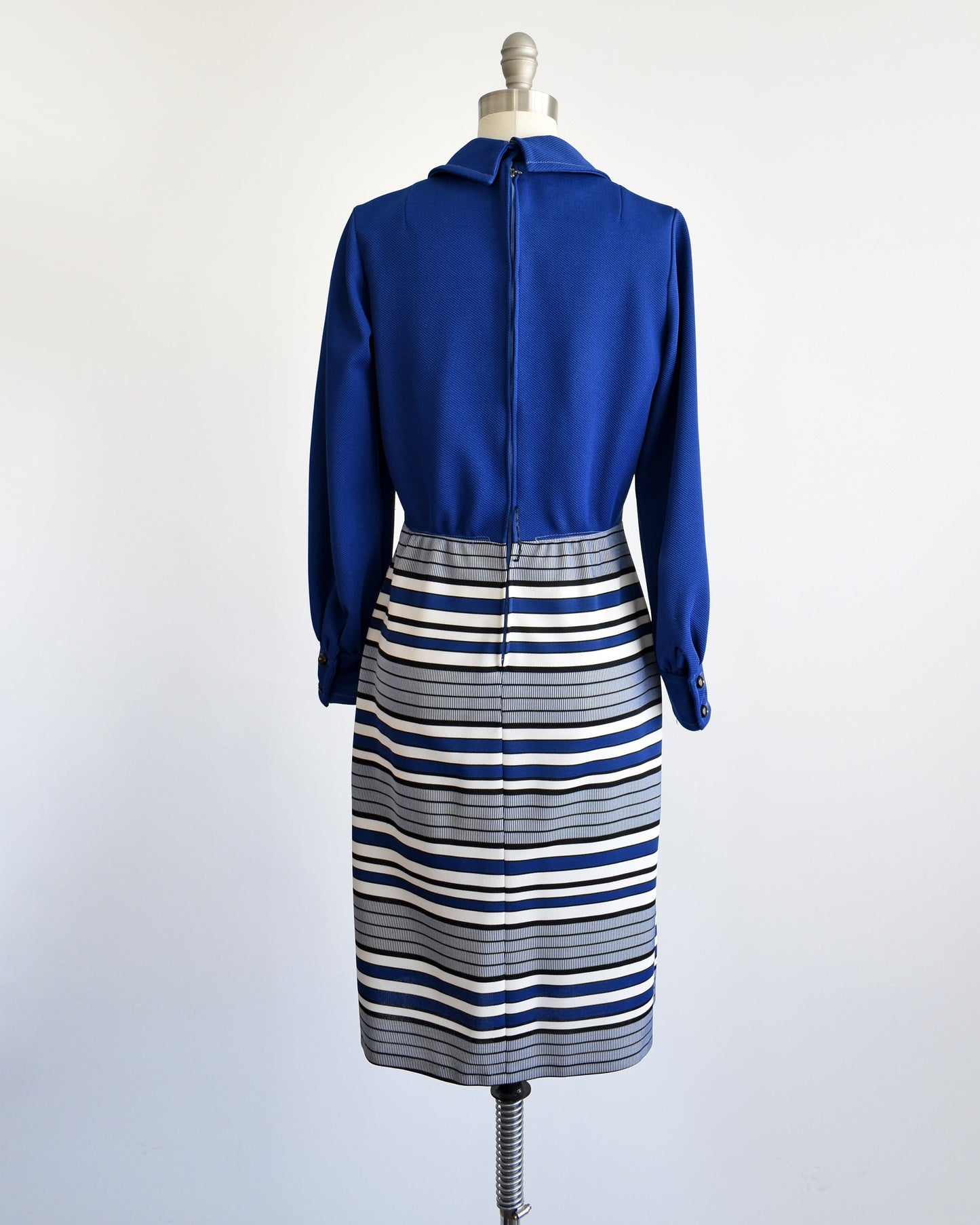 Back view of a vintage 60s mod dress on a dress form. The dress has a dark blue bodice and long sleeves. Blue, black, and white horizontal striped skirt. Zipper up the back