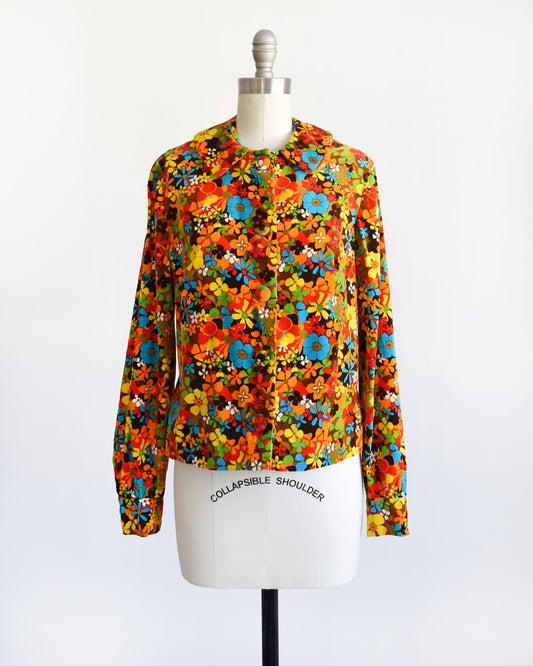 A vintage 70s blouse that has a rainbow flower power print covering the shirt. The blouse is modeled on a dress form