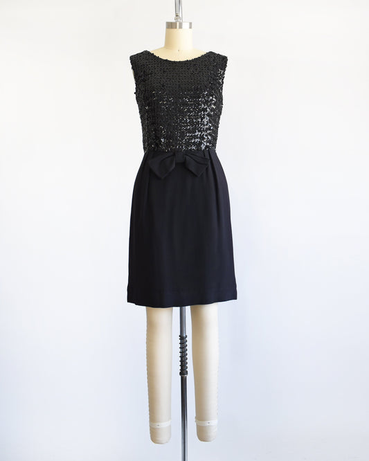 A vintage late 50s early 60s black dress that has a black sequin bodice, a bow at the waist, and a pencil skirt. The dress is modeled on a dress form