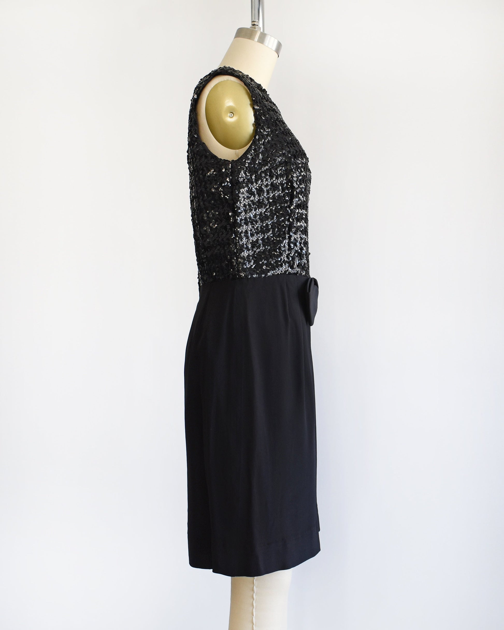 Side view of a vintage late 50s early 60s black dress that has a black sequin bodice, a bow at the waist, and a pencil skirt. The dress is modeled on a dress form