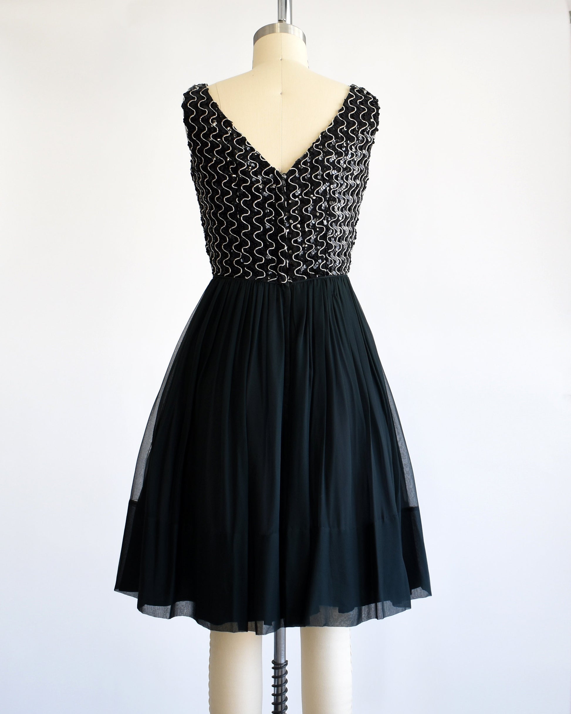 Back view of a vintage 60s dress with a black sequin bodice with silver vertical squiggles. The skirt is a layered with black chiffon on top. The dress is modeled on a dress form.