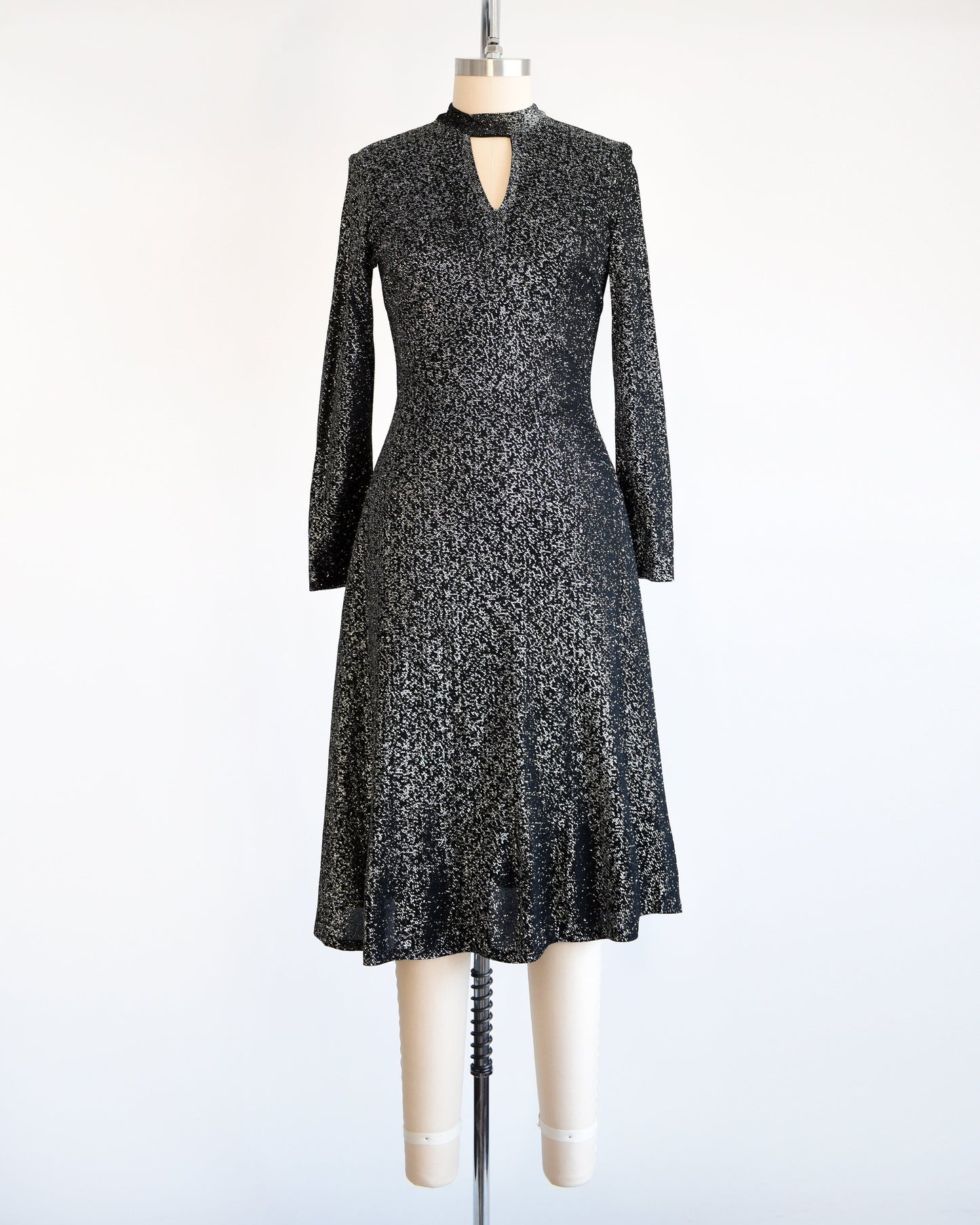 A vintage 70s black and silver metallic dress that has an upside down triangle cutout under the front of the collar, long sleeves, and a knee length skirt. The dress is modeled on a dress form.