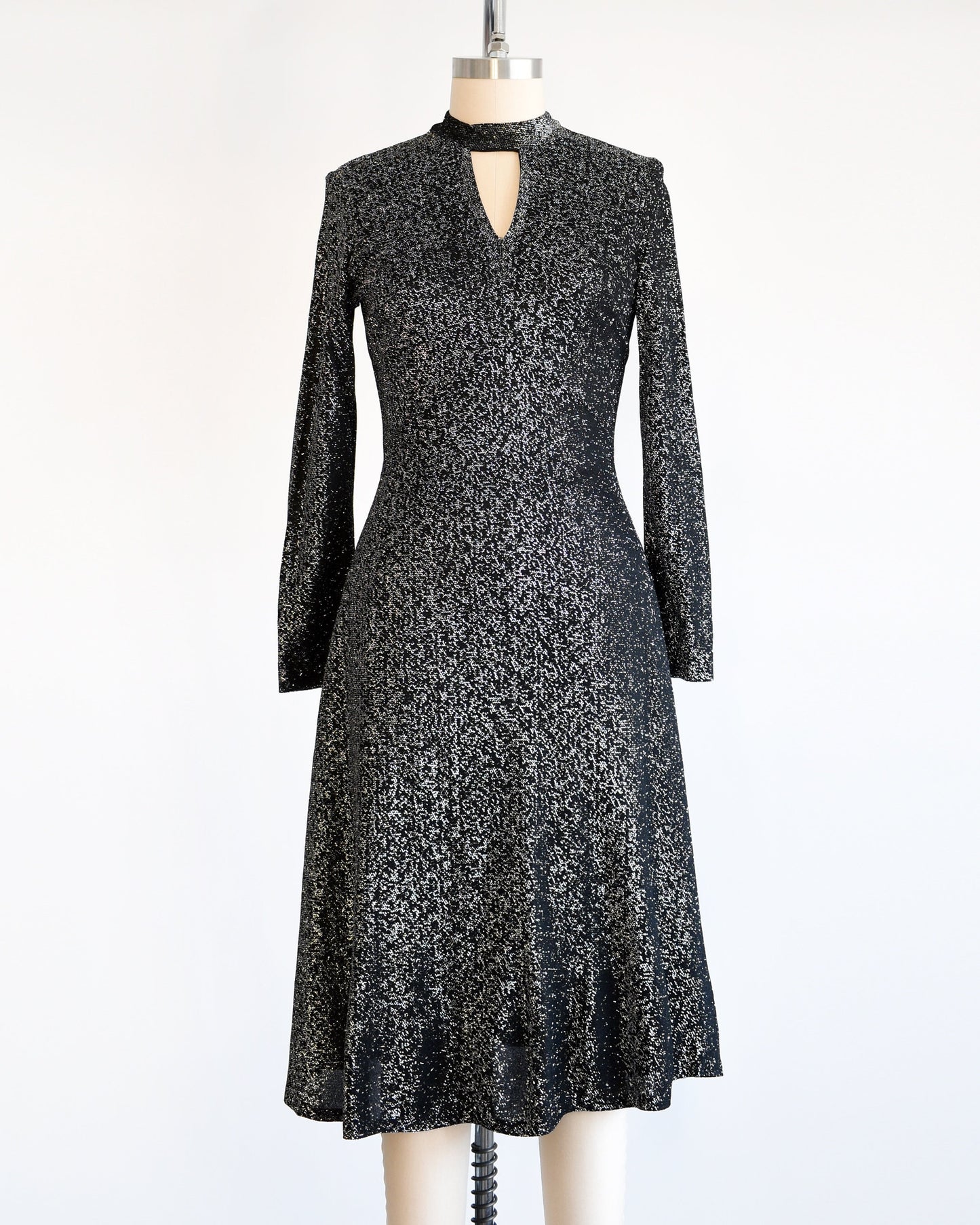 A vintage 70s black and silver metallic dress that has an upside down triangle cutout under the front of the collar, long sleeves, and a knee length skirt. The dress is modeled on a dress form.