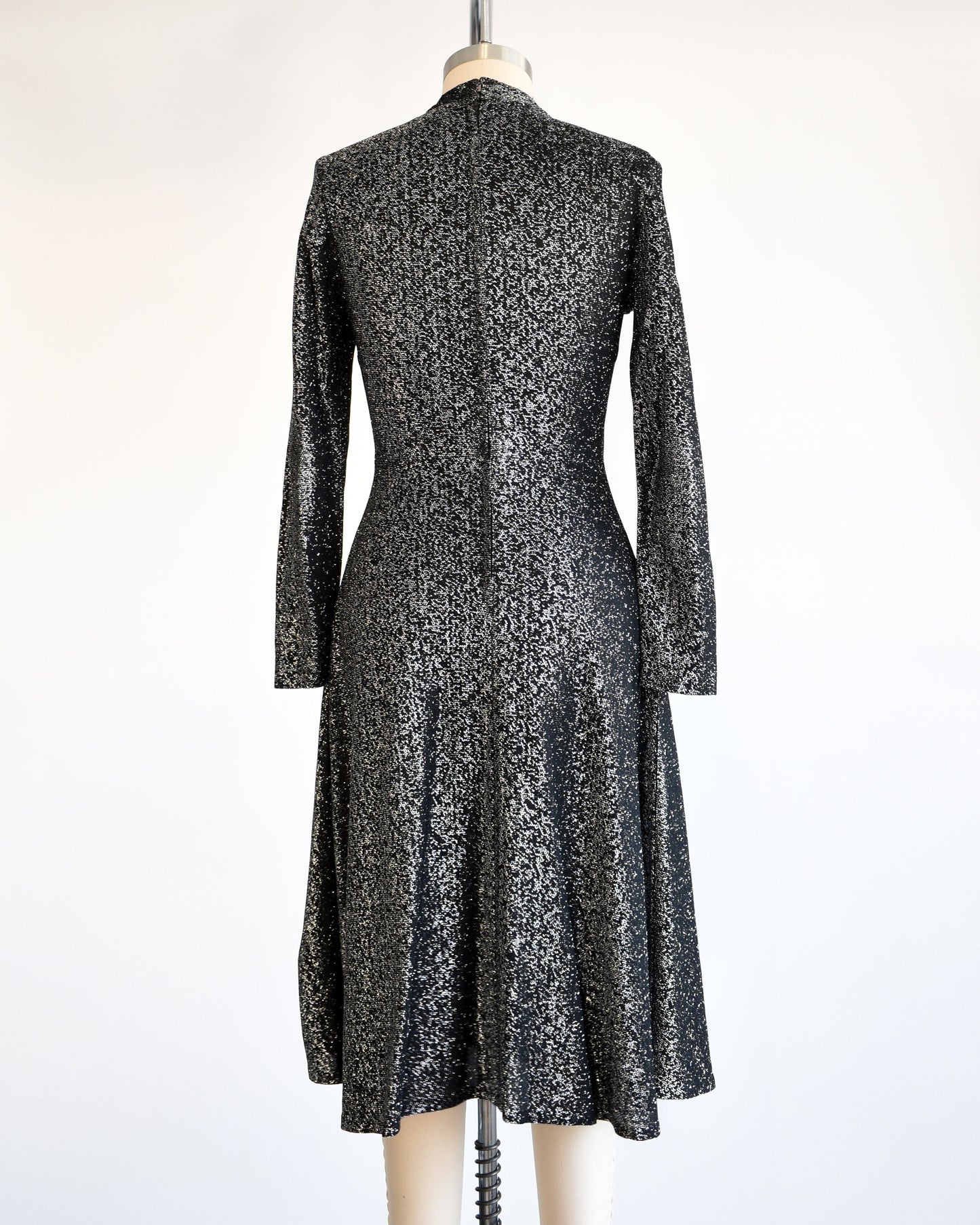 Back view of a vintage 70s black and silver metallic dress that has long sleeves, a zipper up the back, and a knee length skirt. The dress is modeled on a dress form.