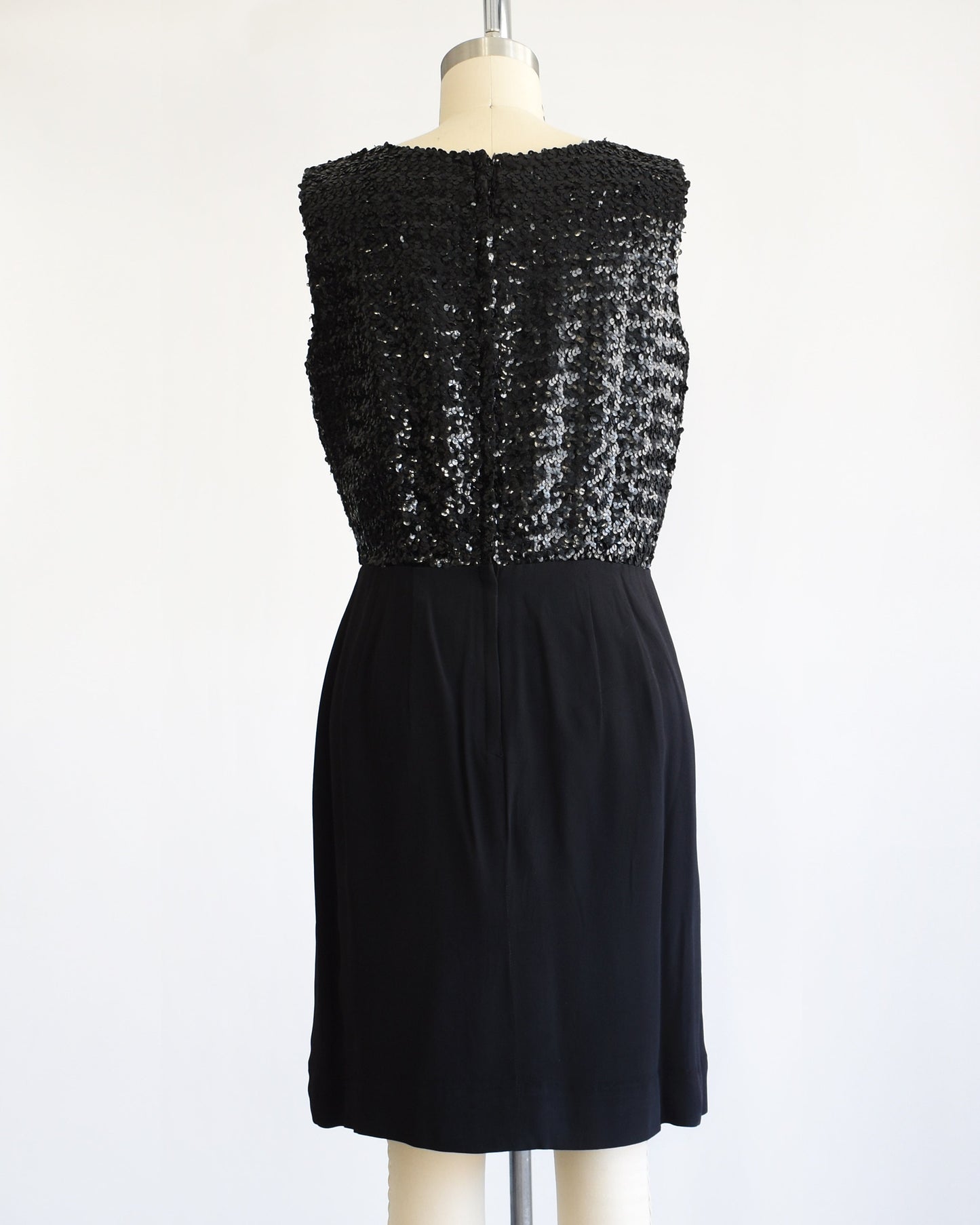 Back view of a vintage late 50s early 60s black dress that has a black sequin bodice and a pencil skirt. The dress is modeled on a dress form