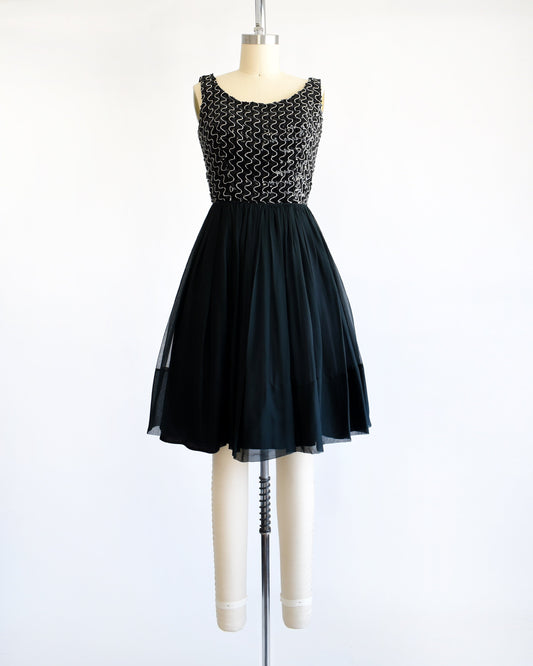 A vintage 60s dress with a black sequin bodice with silver vertical squiggles. The skirt is a layered with black chiffon on top. The dress is modeled on a dress form.