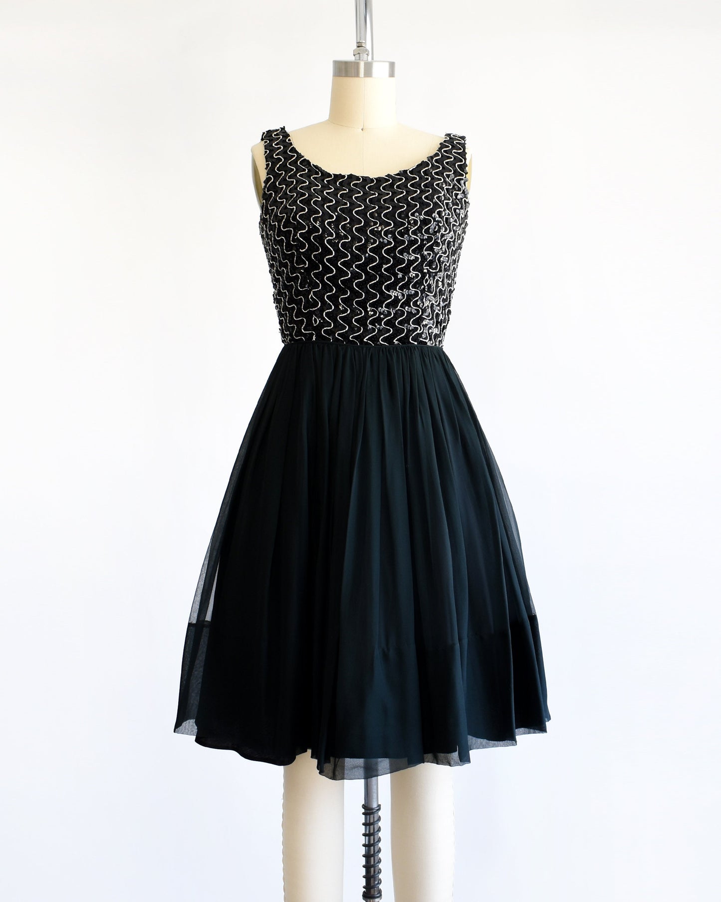 A vintage 60s dress with a black sequin bodice with silver vertical squiggles. The skirt is a layered with black chiffon on top. The dress is modeled on a dress form.