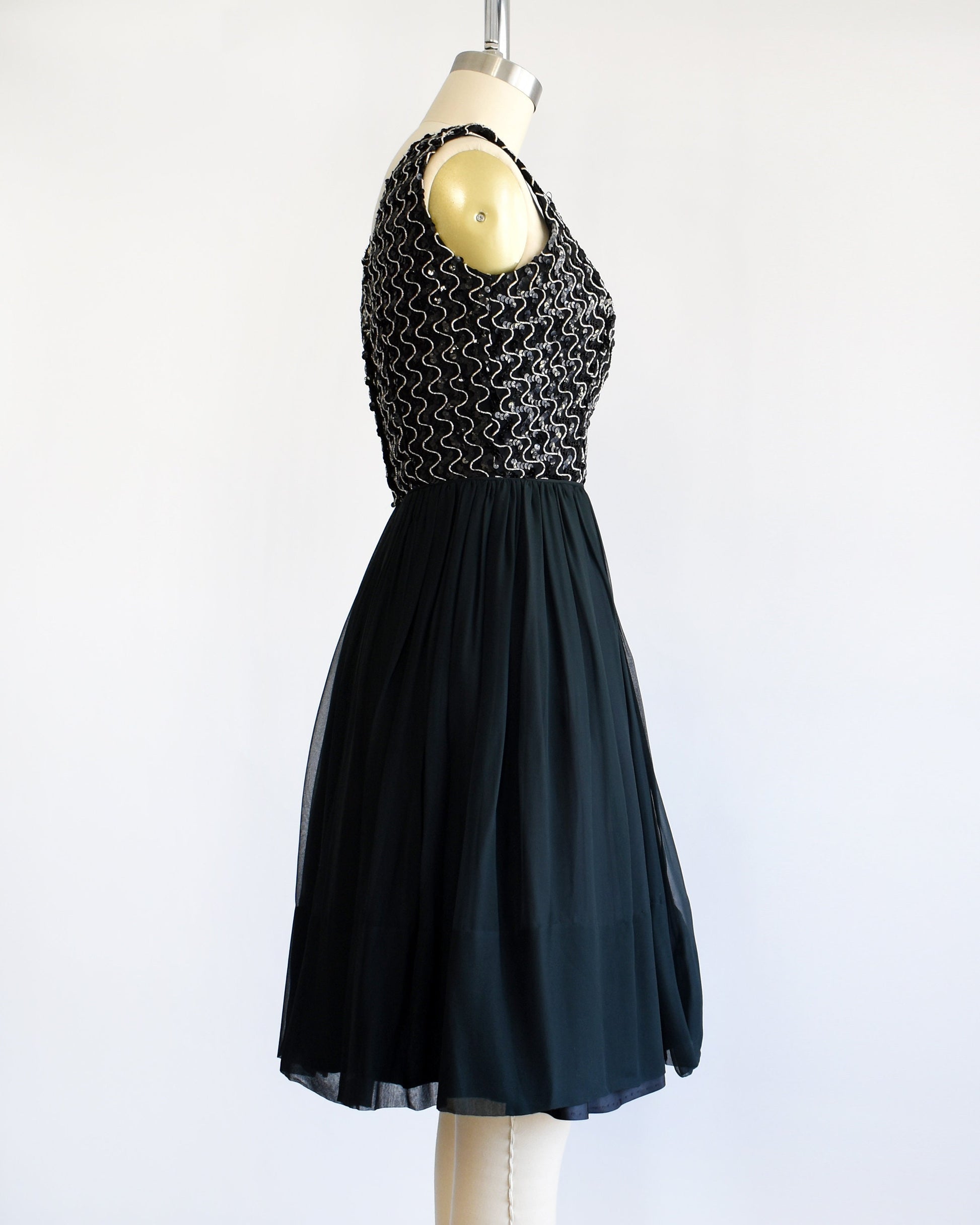 Side view of a vintage 60s dress with a black sequin bodice with silver vertical squiggles. The skirt is a layered with black chiffon on top. The dress is modeled on a dress form.