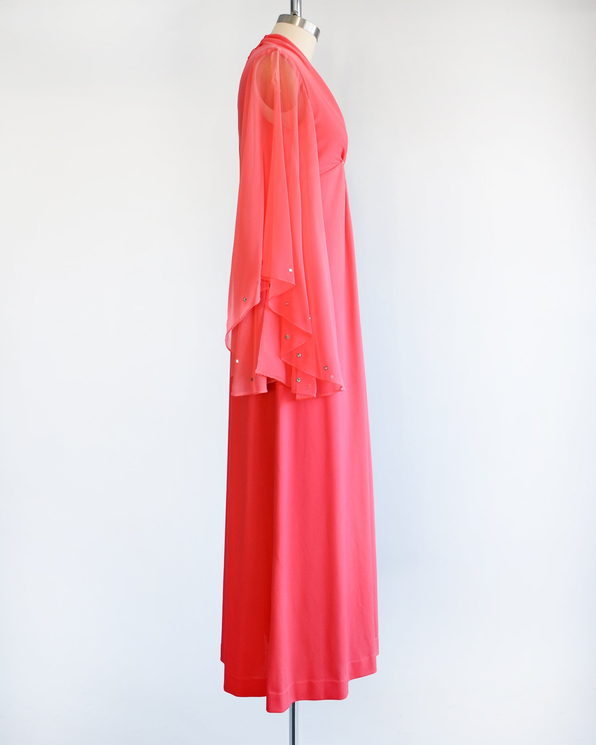 Side view of a vintage 70s coral pink maxi dress that has a v-neckline, rhinestones on the waist, and semi-sheer long angel sleeves with matching rhinestone trim. The dress is modeled on a dress form.