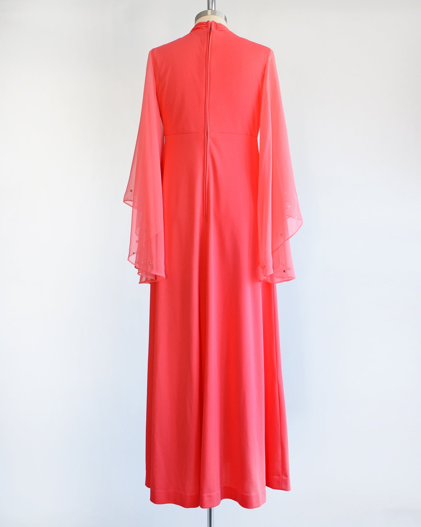 Back view of a vintage 70s coral pink maxi dress that has semi-sheer long angel sleeves with matching rhinestone trim. The dress is modeled on a dress form.
