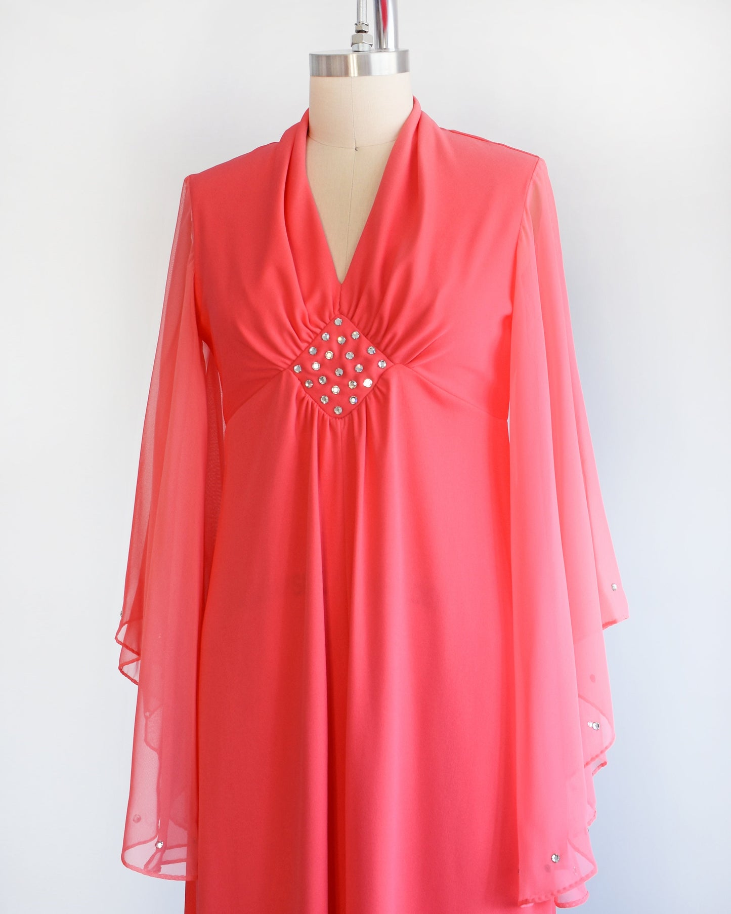 Side front view of a vintage 70s coral pink maxi dress that has a v-neckline, rhinestones on the waist, and semi-sheer long angel sleeves with matching rhinestone trim. The dress is modeled on a dress form.