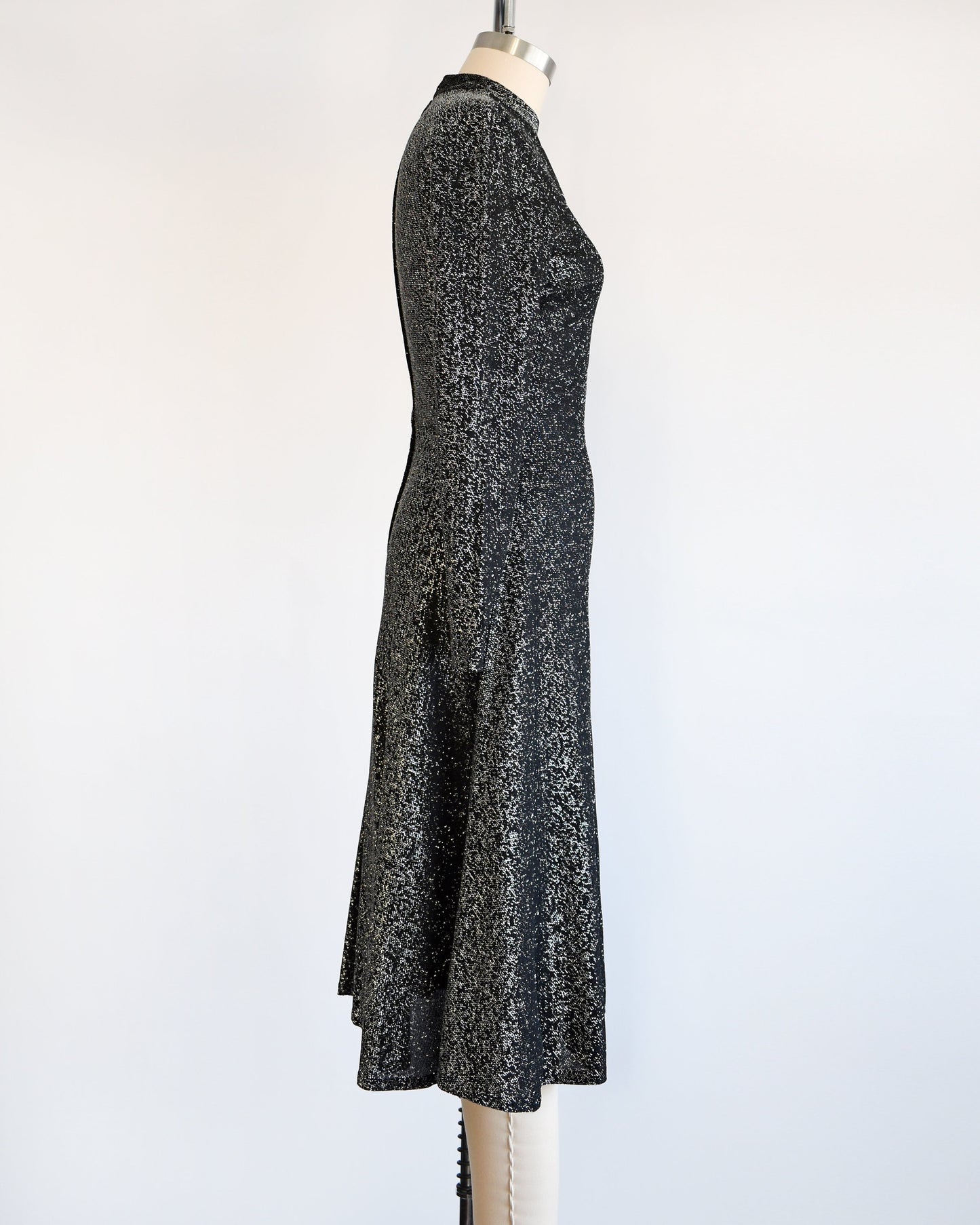 Side view of a vintage 70s black and silver metallic dress that has long sleeves and a knee length skirt. The dress is modeled on a dress form.