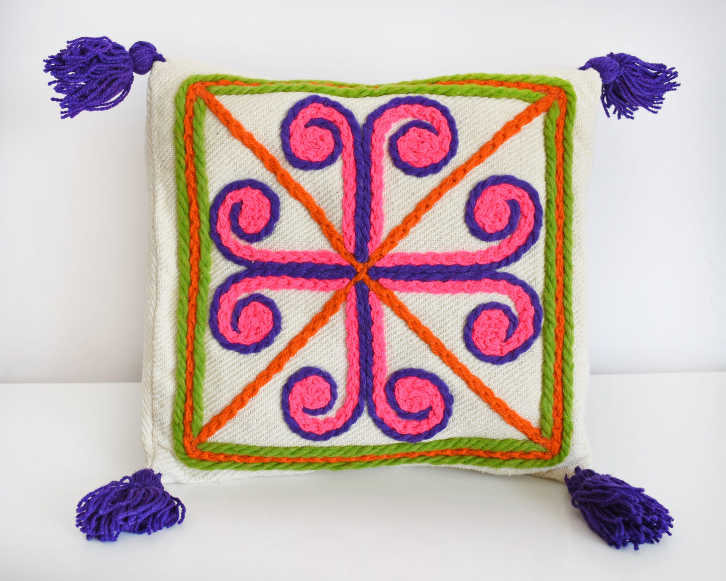 A vintage crewel embroidered pillow that has a curly cross with purple tassels.