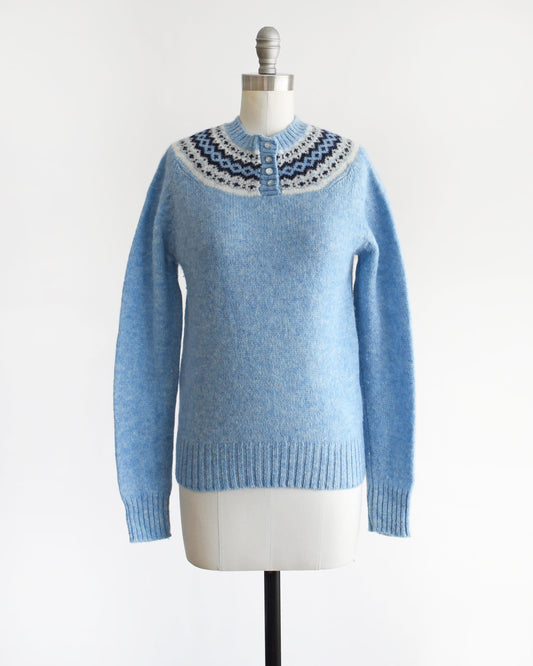 A vintage 80s blue sweater with a fair isle pattern around the collar and 4 buttons down the front. The sweater is modeled on a dress form.