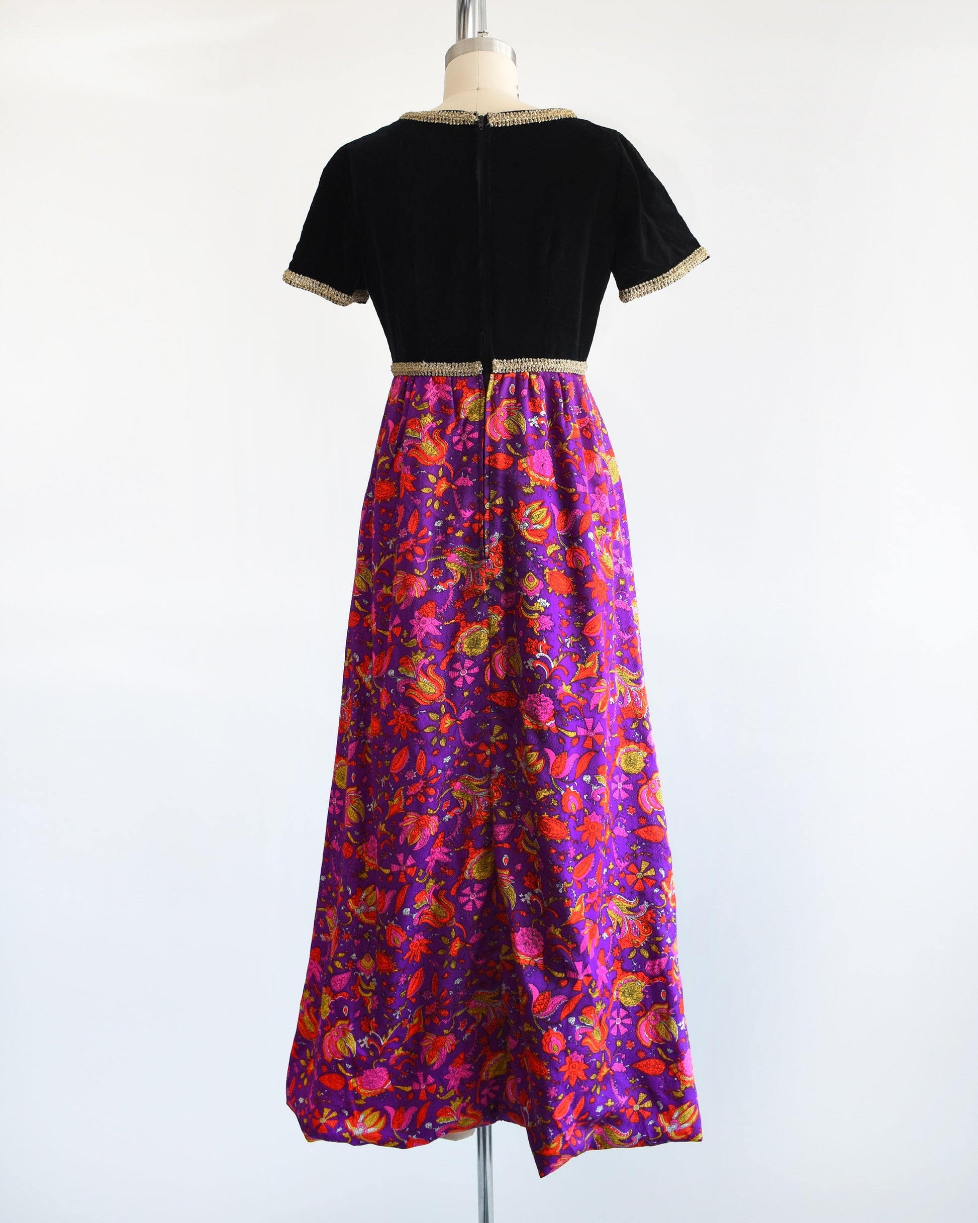 Back view of a vintage 60s/70s maxi dress that has a black velvet bodice with gold metallic ribbon trim, along with a purple skirt with pink, orange, yellow, and white floral print.