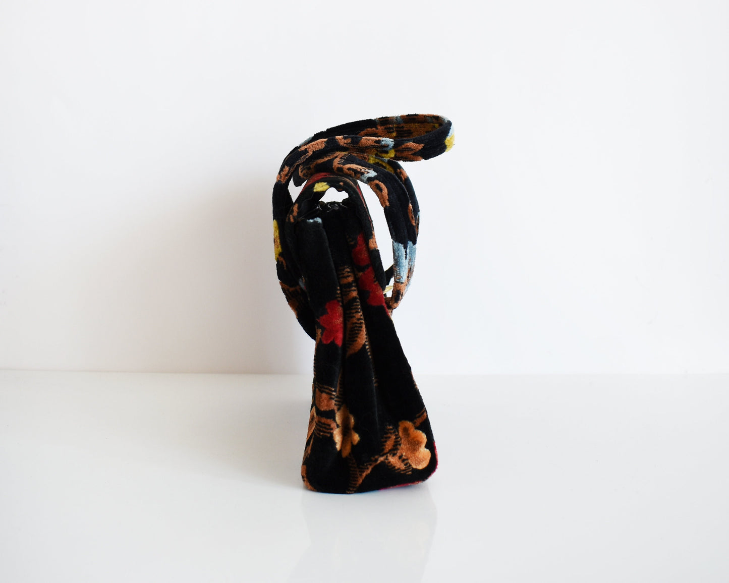 Side view of a vintage black floral velvet chenille handbag on a white table. The flowers are red, blue, yellow and have brown stems and leaves.