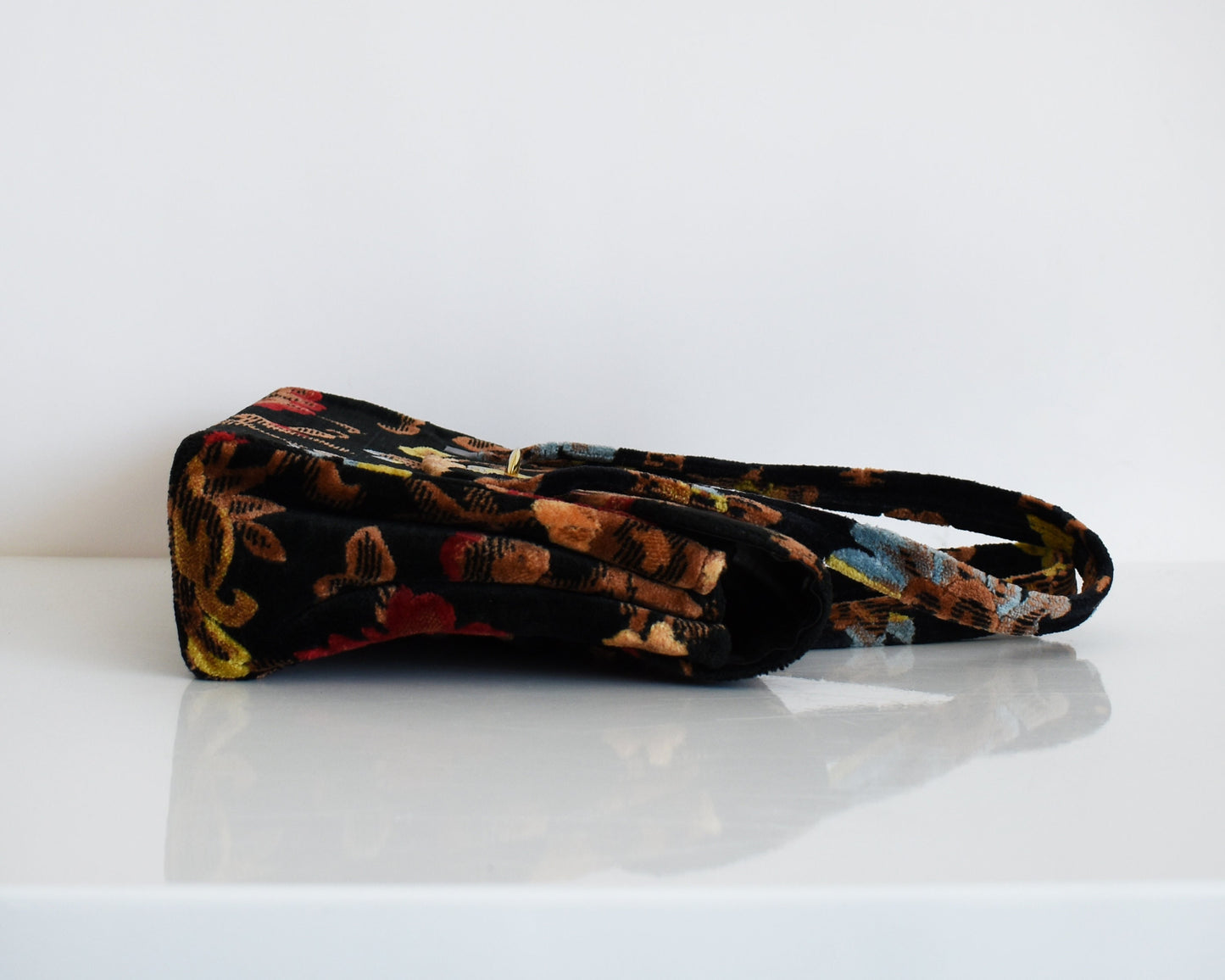 A vintage black floral velvet chenille handbag with gold tone hardware laying on a table. The flowers are red, blue, yellow and have brown stems and leaves.
