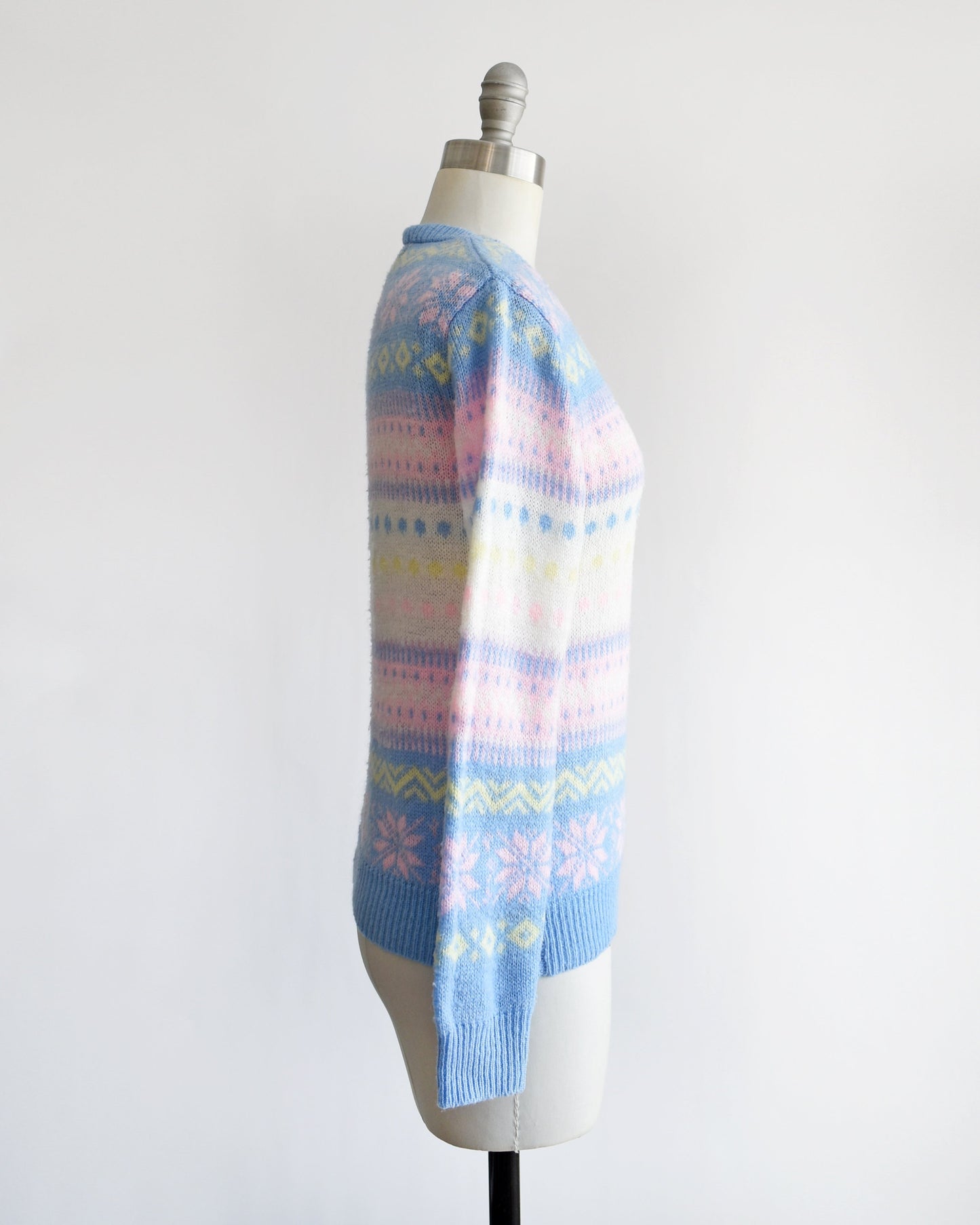 Side view of a vintage 80s pastel striped sweater thats blue, pink, yellow, and white, and at has a snowflake and geometric stripe print on a dress form.