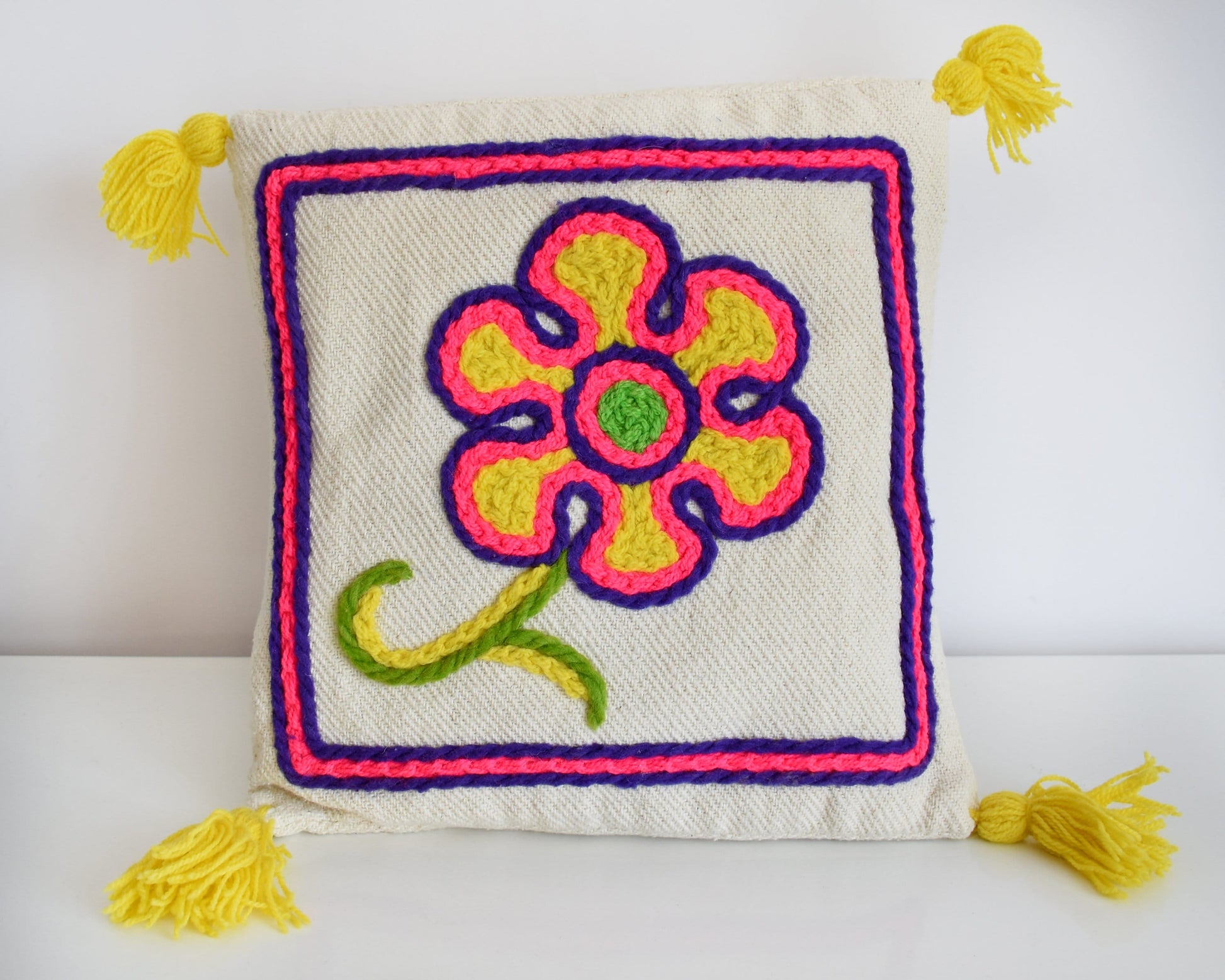 A vintage crewel embroidered floral pillow with yellow tassels on a table.