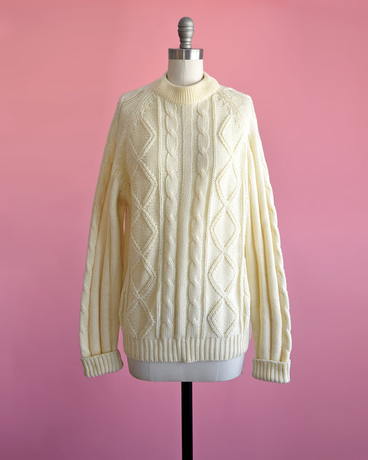 A vintage 70s cream cable knit sweater on a dress form set against a pink background