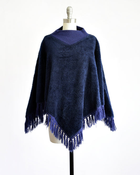A vintage 70s dark blue fuzzy fleece poncho with a ribbed collar and fringe along the hem on a dress form.