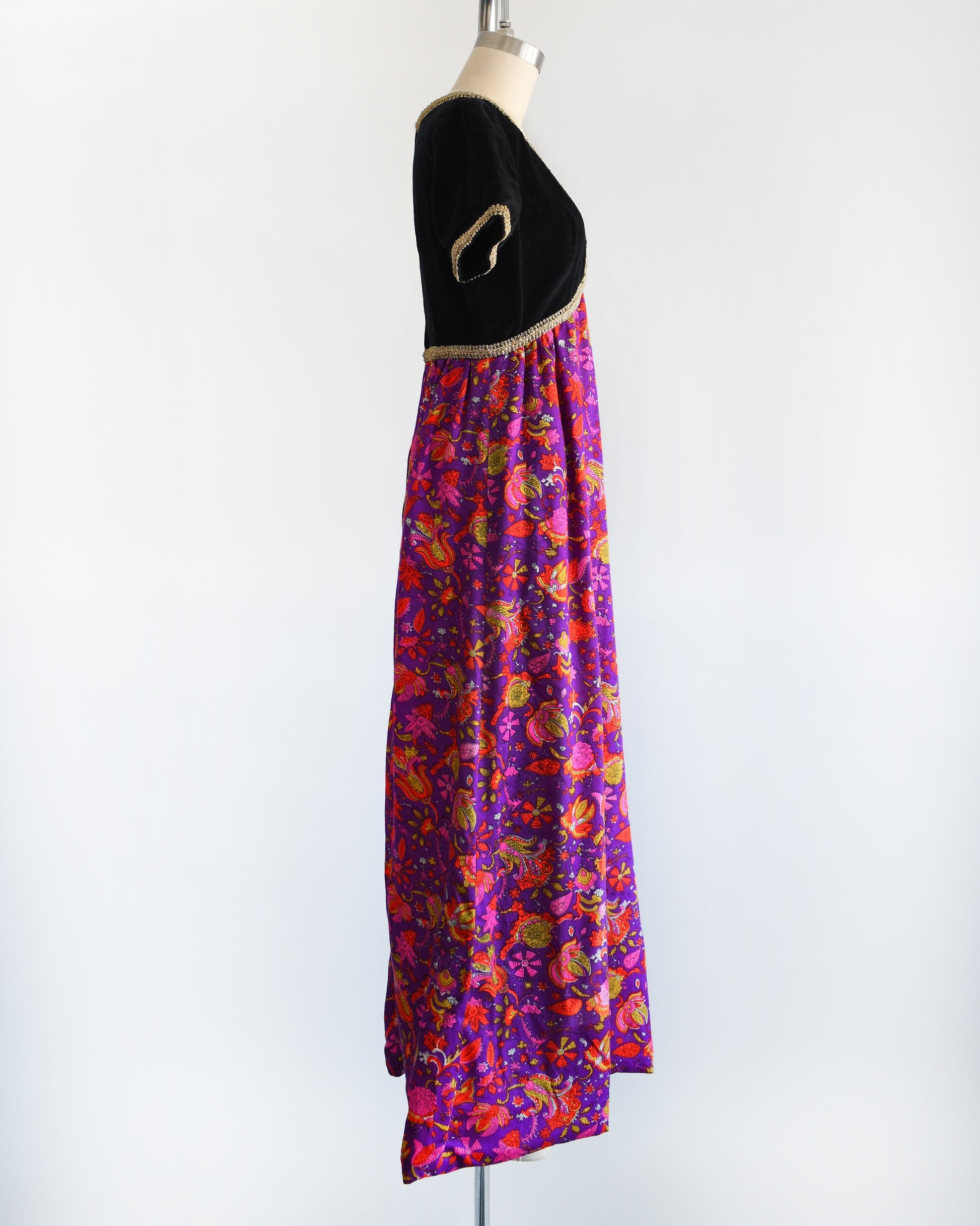 Side view of a vintage 60s/70s maxi dress that has a black velvet bodice with gold metallic ribbon trim, along with a purple skirt with pink, orange, yellow, and white floral print.