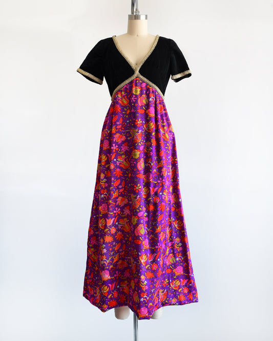 A vintage 60s/70s maxi dress that has a black velvet bodice with gold metallic ribbon trim, along with a purple skirt with pink, orange, yellow, and white floral print.