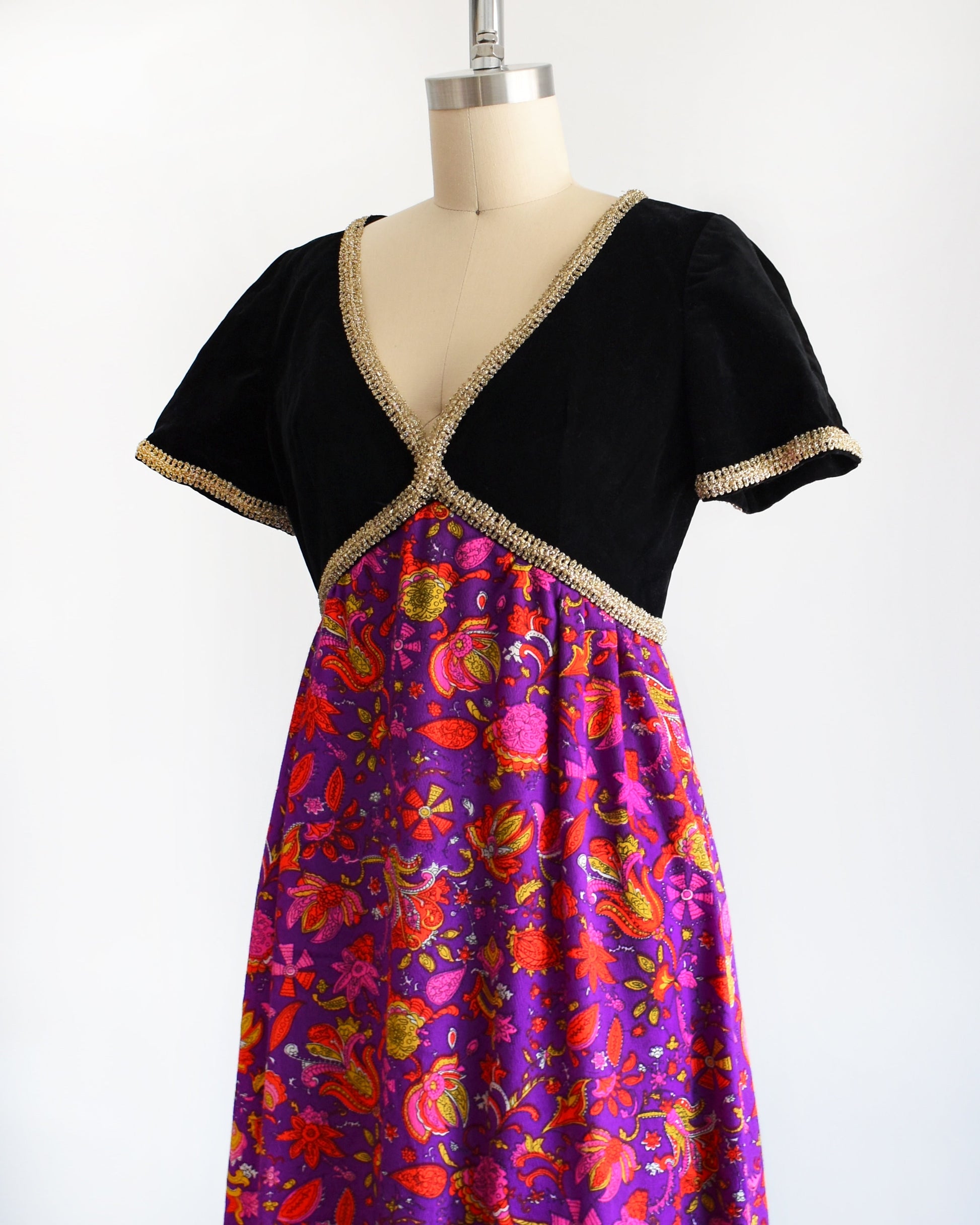Side front view of a vintage 60s/70s maxi dress that has a black velvet bodice with gold metallic ribbon trim, along with a purple skirt with pink, orange, yellow, and white floral print.