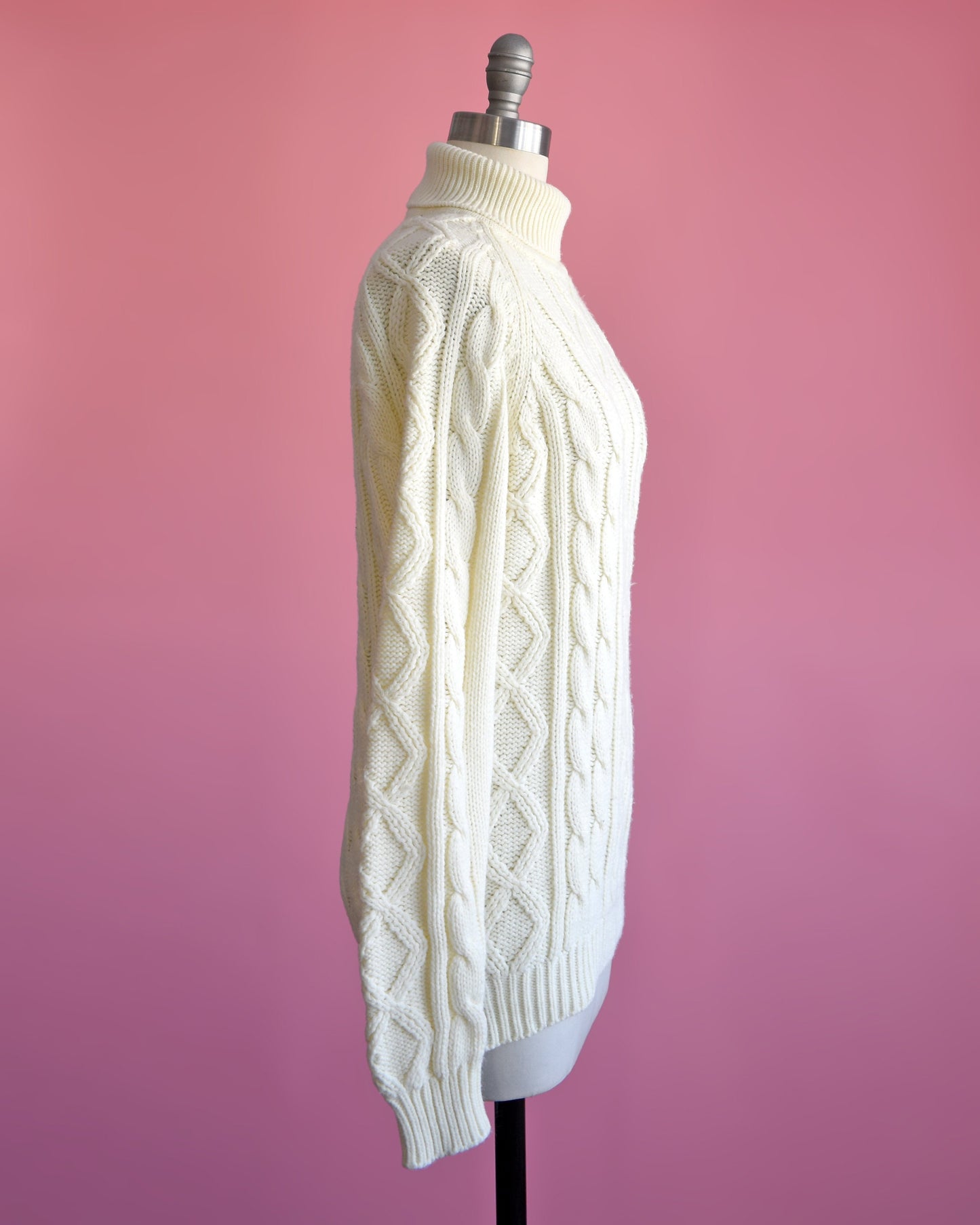side view of a vintage 70s cream cable knit turtleneck sweater on a dress form set against a pink background