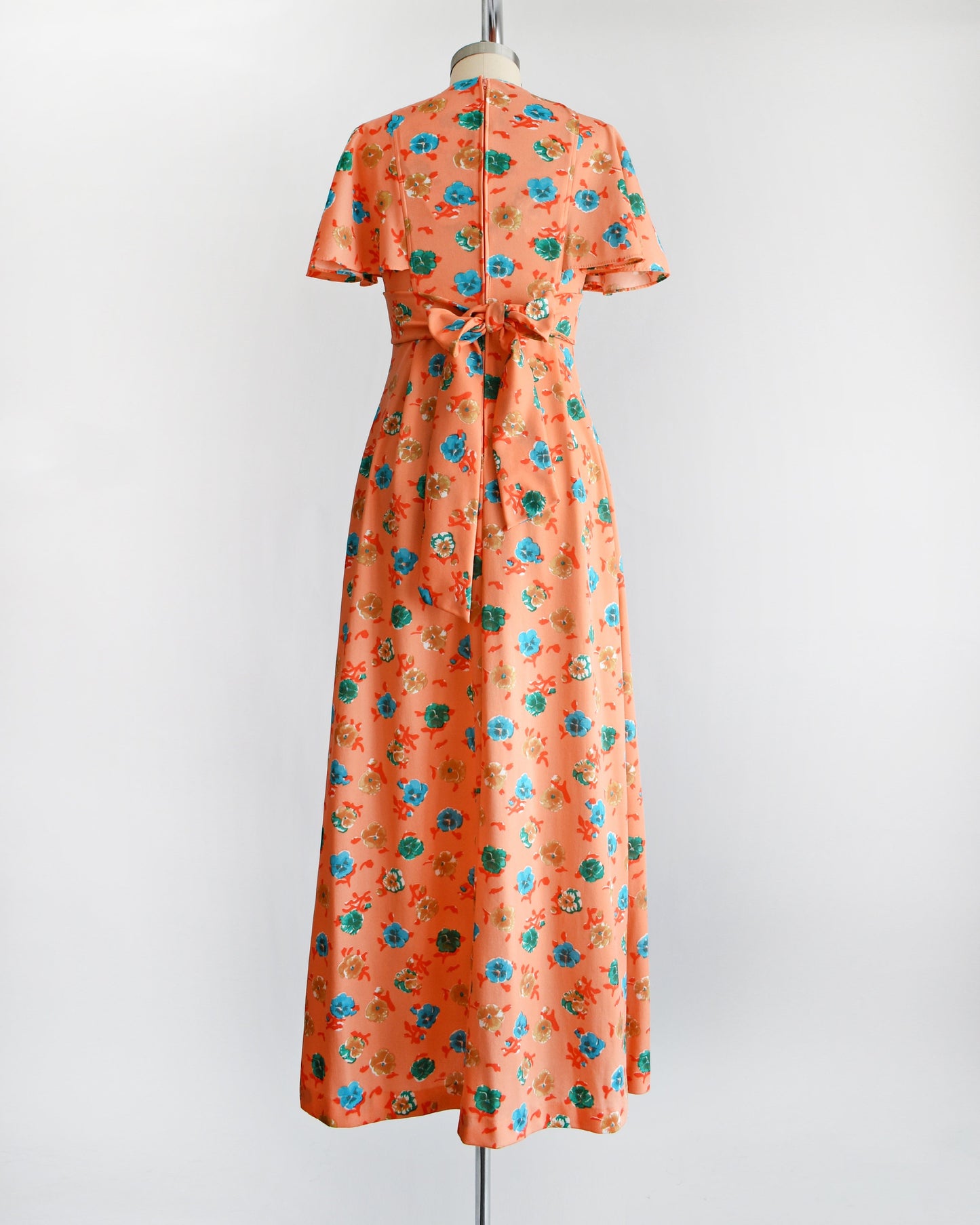 Back view of a vintage 70s orange maxi dress with a blue, green, and brown floral print, along with flutter sleeves, a v-neckline, and empire waist that ties in the back. The dress is modeled on a dress form.