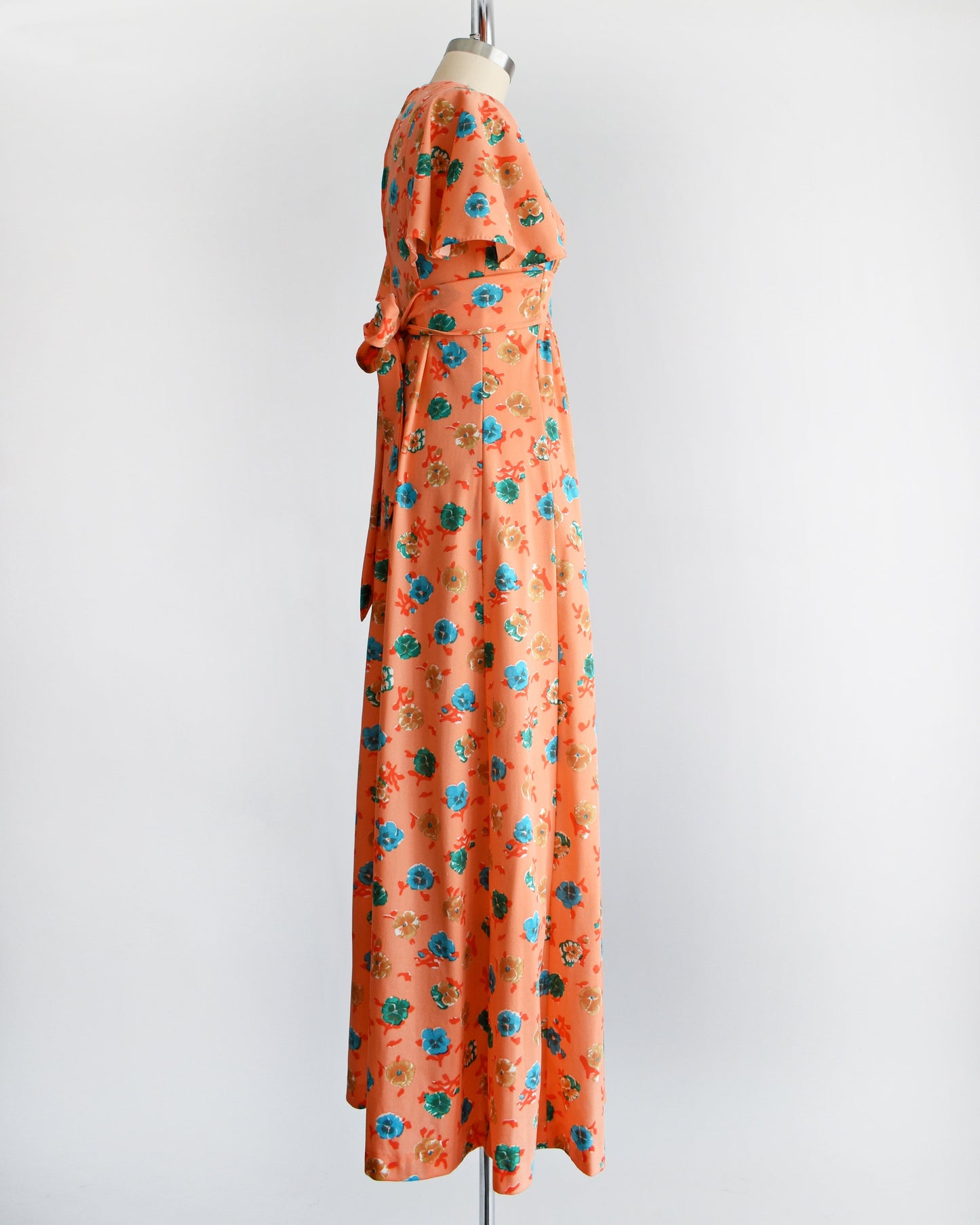 Side view of a vintage 70s orange maxi dress with a blue, green, and brown floral print, along with flutter sleeves, a v-neckline, and empire waist. The dress is modeled on a dress form.