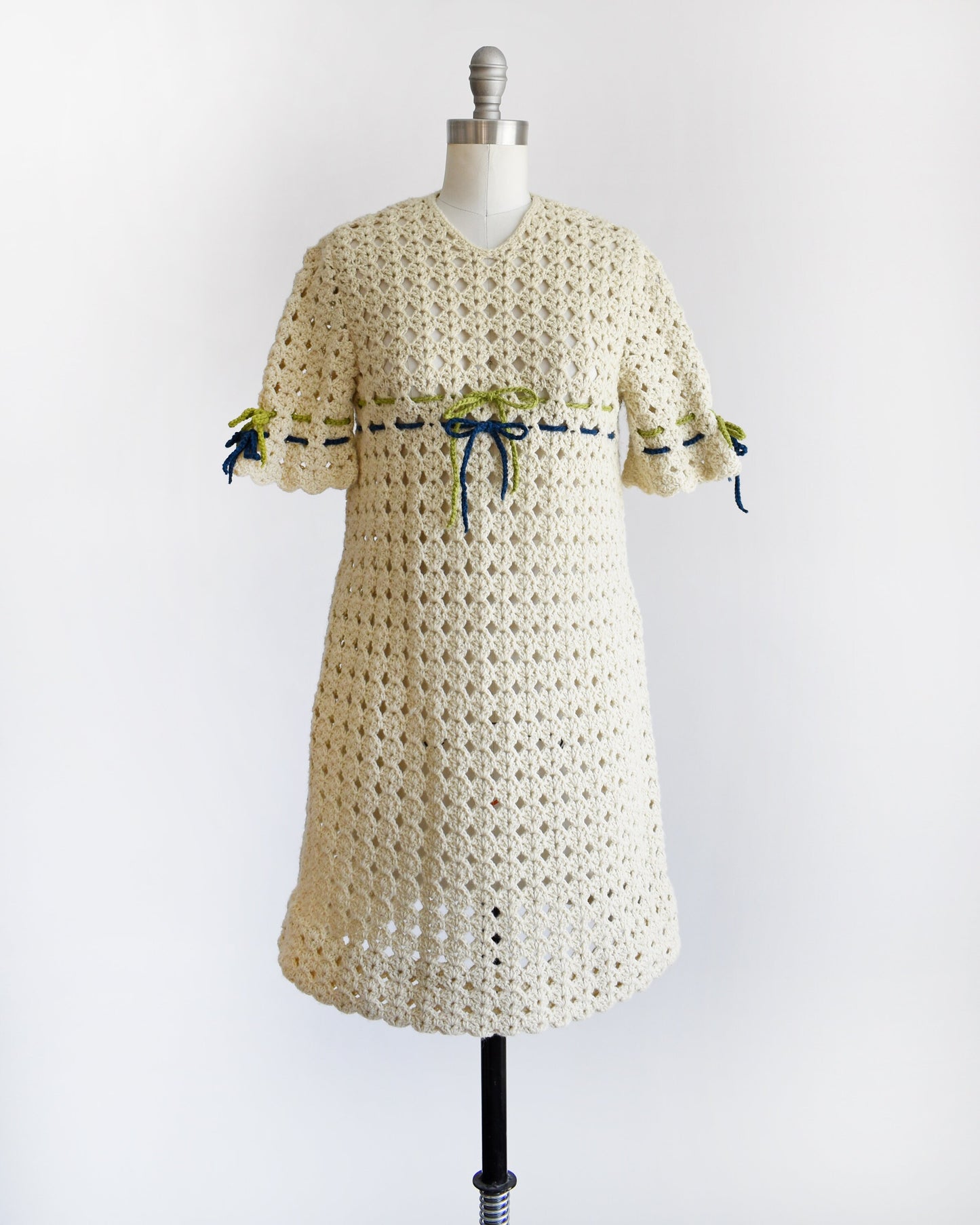 A vintage 70s crochet mini dress with a green and blue woven stripe on the front that ties into a bow, along with matching stripes and bows on the sleeves. The dress is on a dress form.
