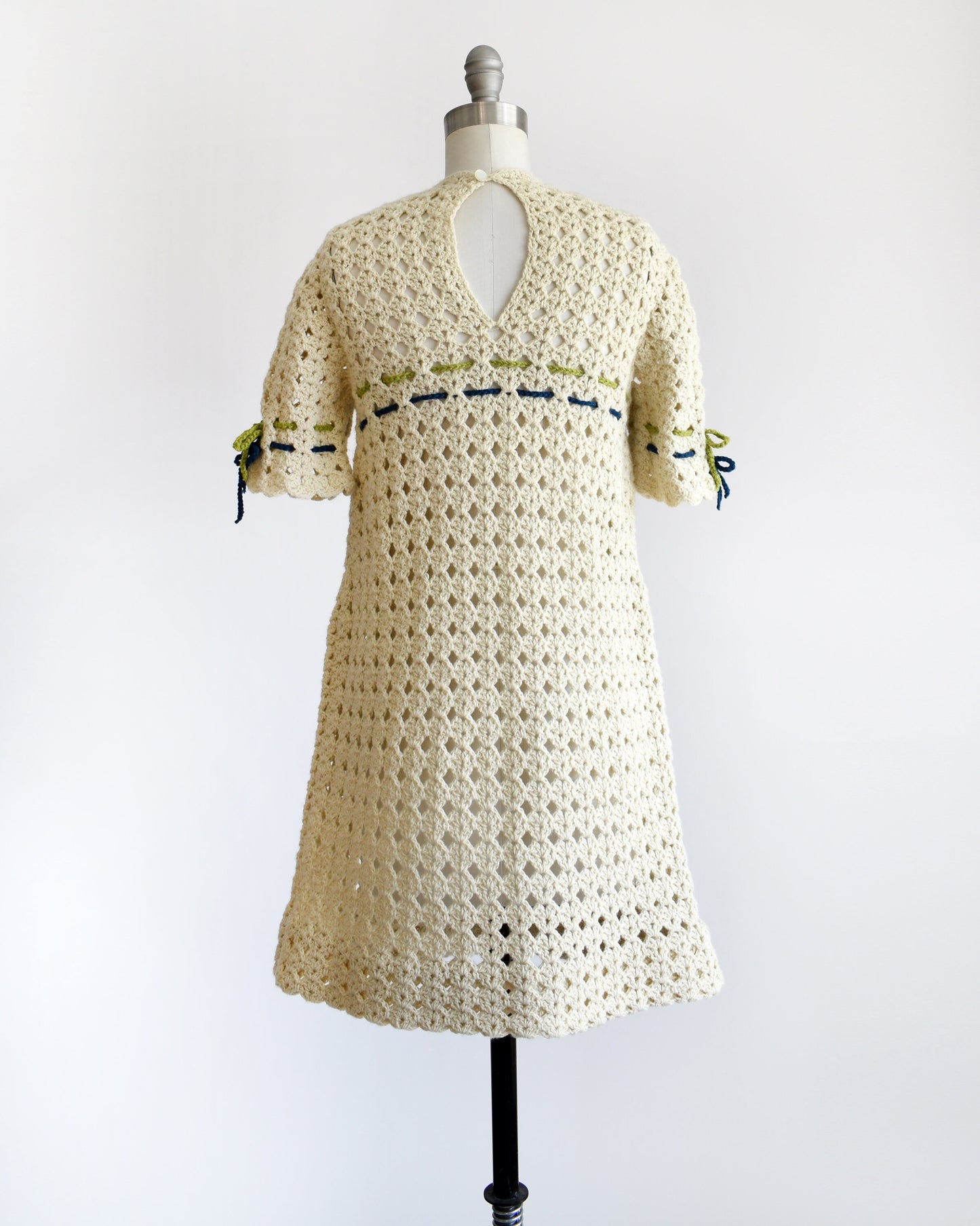 Back view of  vintage 70s crochet mini dress with a green and blue woven stripe on the front that ties into a bow, along with matching stripes and bows on the sleeves. One button at the back of the neck which has a keyhole underneath