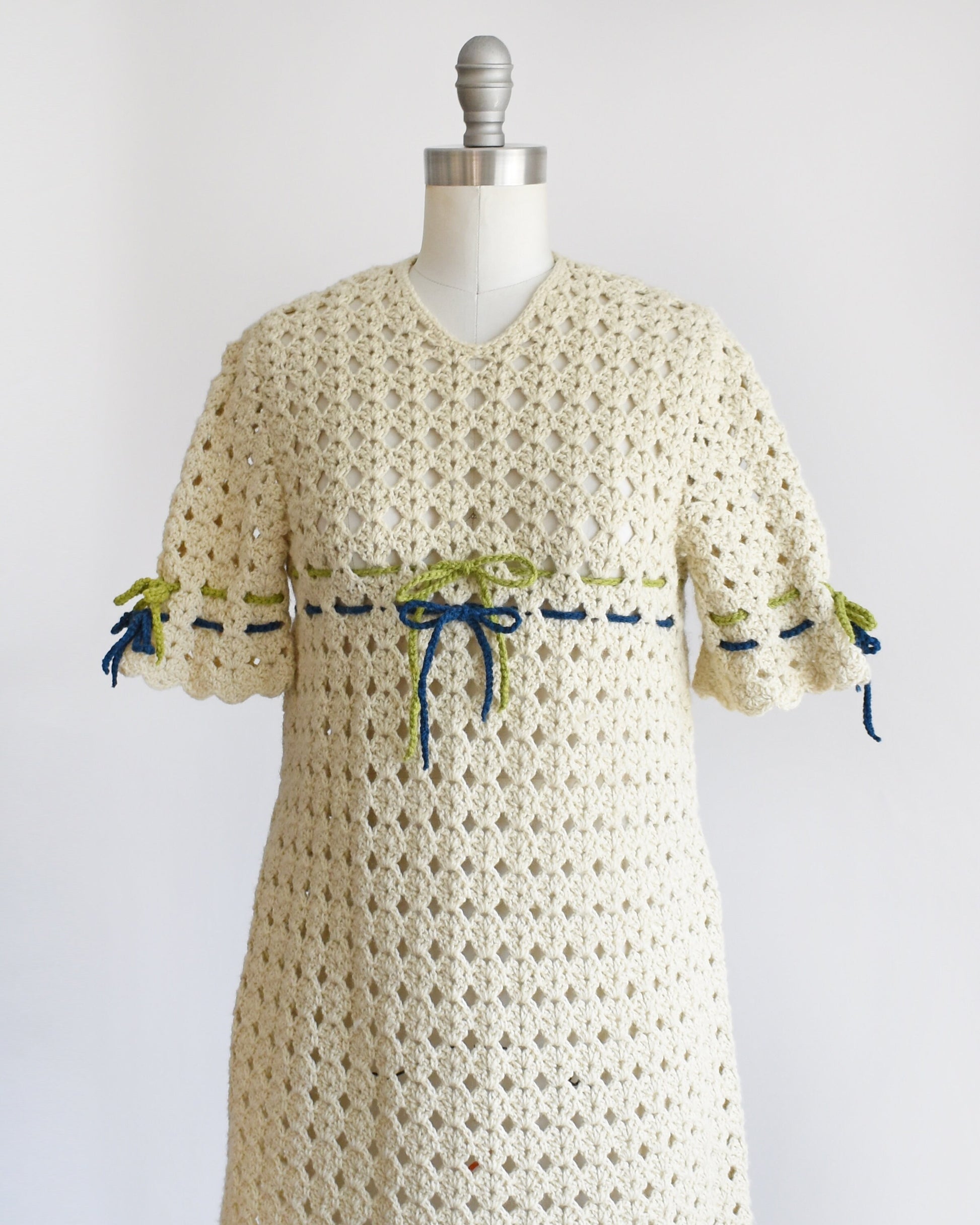 Side front view of a vintage 70s crochet mini dress with a green and blue woven stripe on the front that ties into a bow, along with matching stripes and bows on the sleeves. The dress is on a dress form.