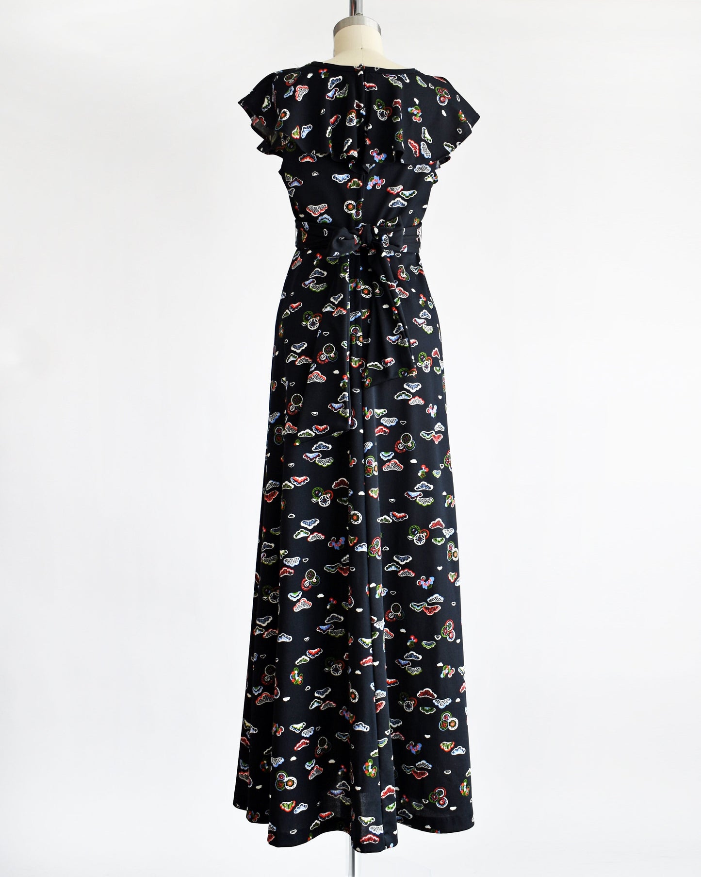 Back view of a vintage 70s flutter sleeve black dress with a multicolored abstract print of different shapes and circles. The dress has ties on the sides that tie on the back and is modeled on a dress form.