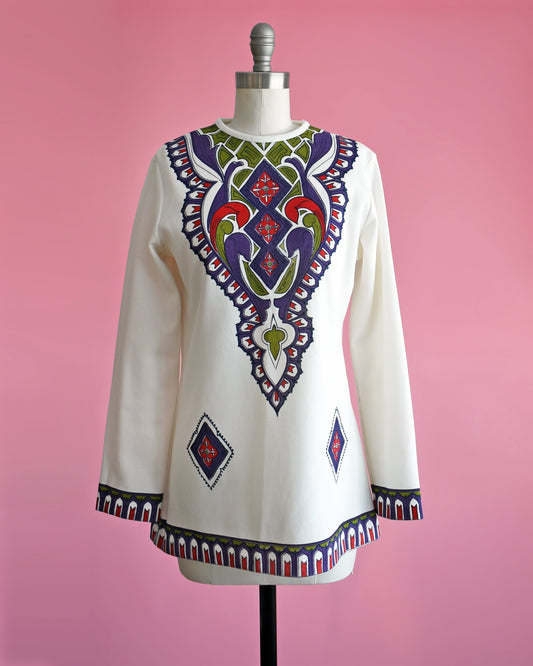 A vintage 60s/70s white tunic with a purple, green, red, and black psychedelic print on the front. Matching print along the hem and long sleeve cuffs. The top is on a dress form.
