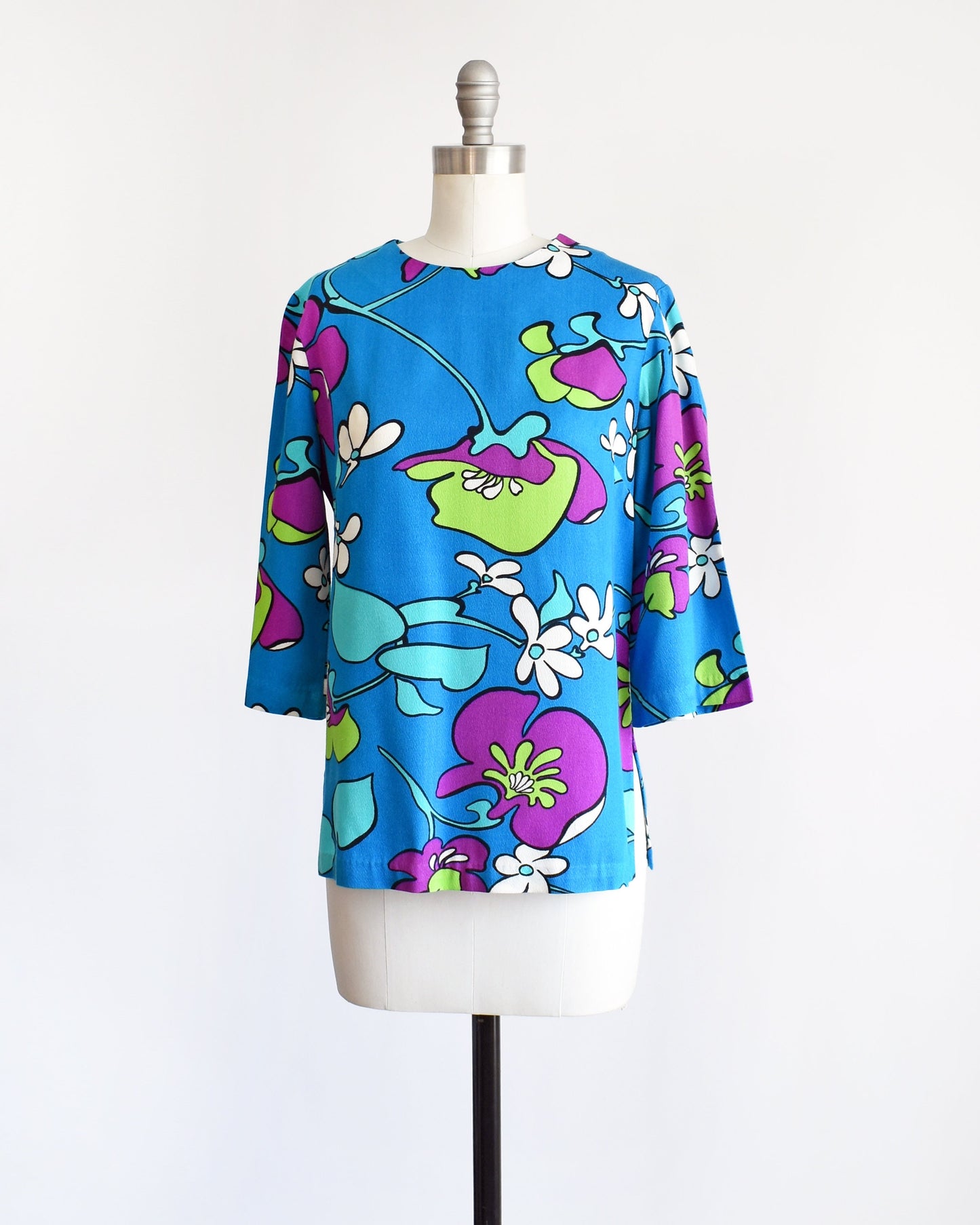 Side front view of a vintage blue blouse with a large purple, green, white, and light blue floral print on a dress form.