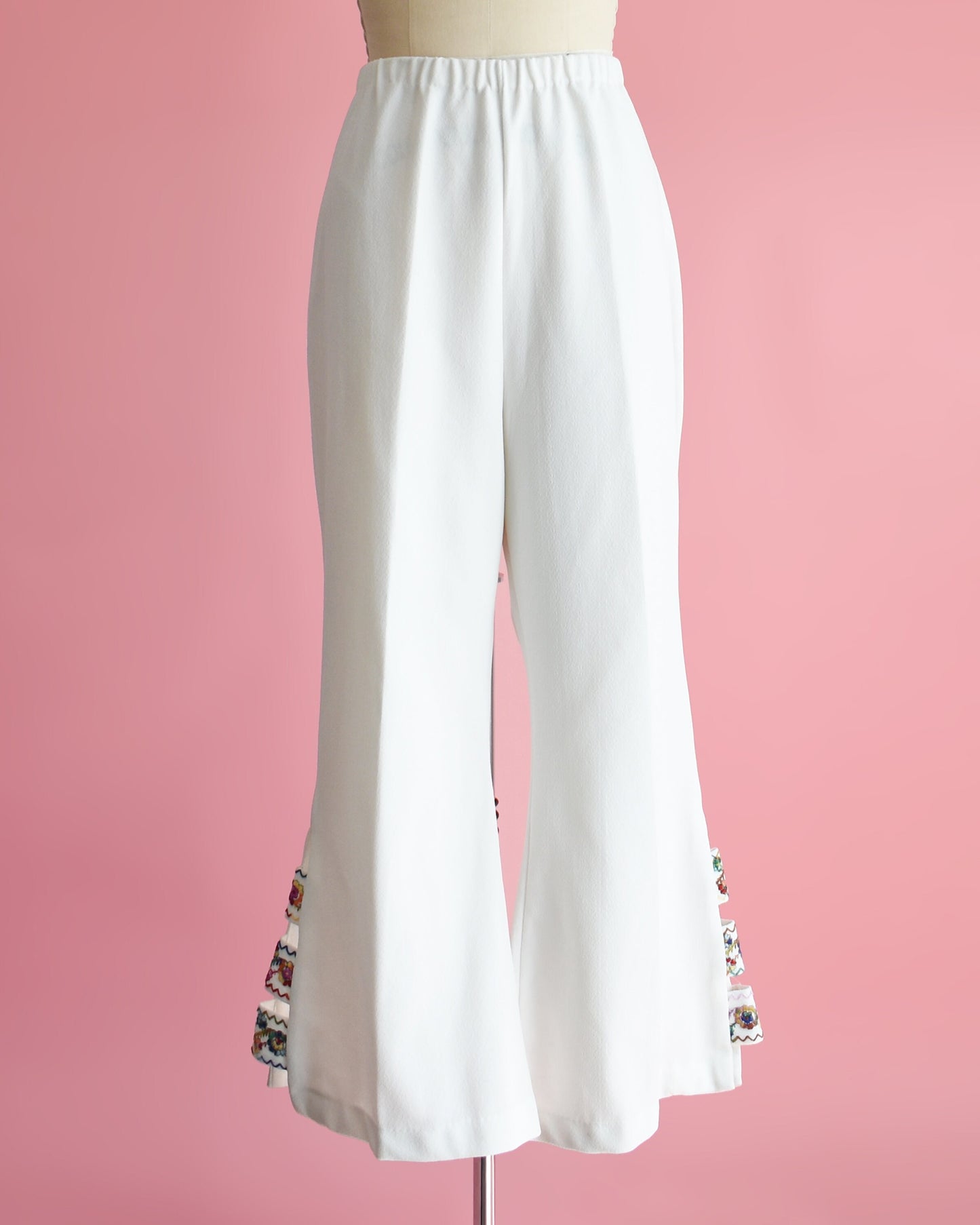 A pair of vintage 70s white bell bottom pants that have embroidered cut out detail on the side of the cuffs. The pants are on a dress form.